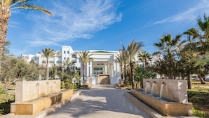 Una entrada de hotel con un camino central, palmeras y una fuente de agua.
