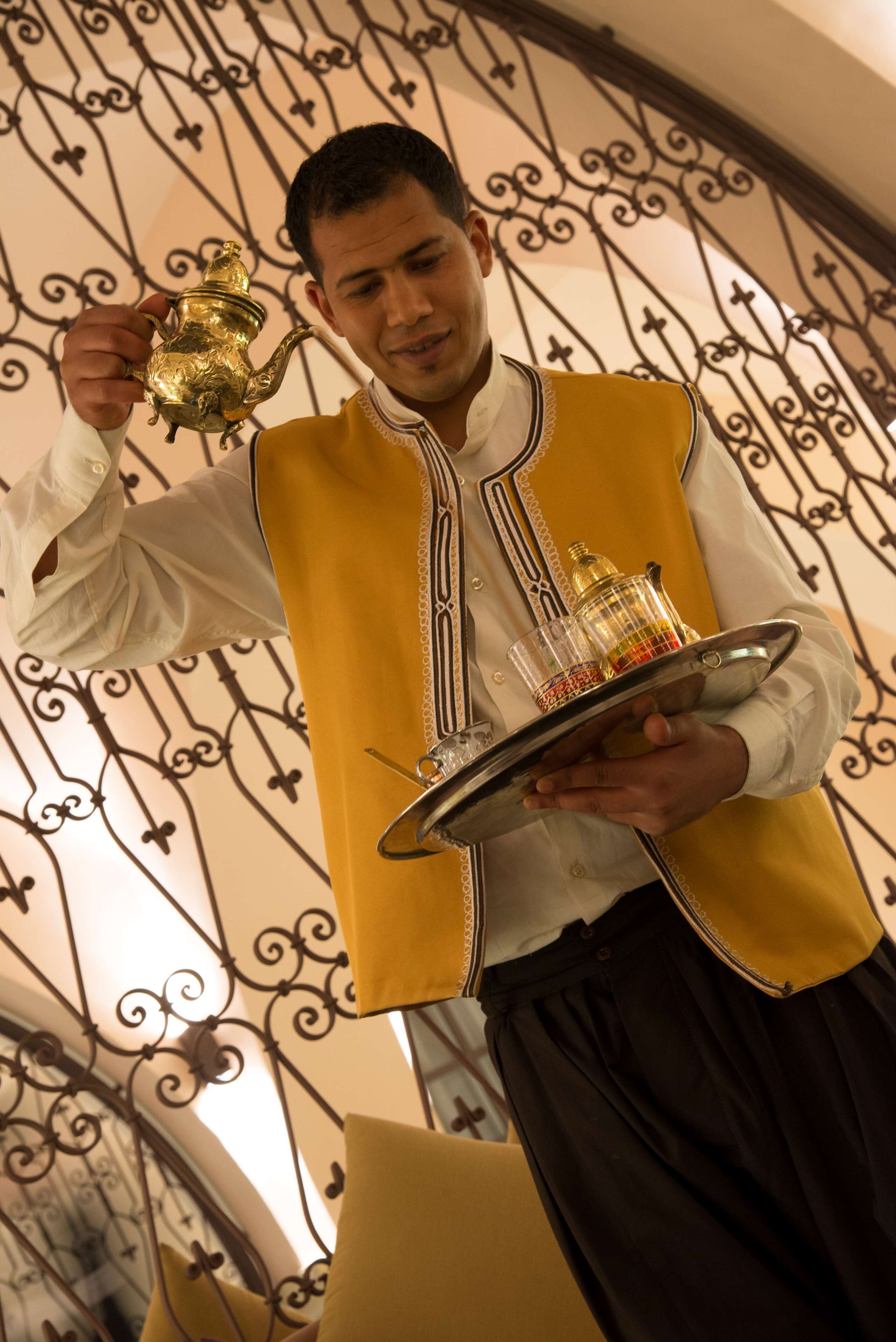 A person in a yellow vest holding a tray with a golden teapot and glasses.
