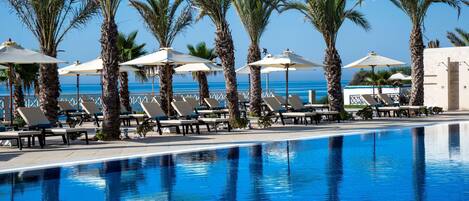 A poolside area with lounge chairs and umbrellas, palm trees, and a view of the ocean.