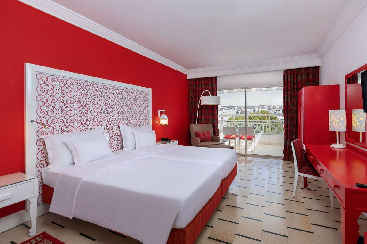 A hotel room with a large bed, a desk, a chair, a TV, and a balcony with a view.