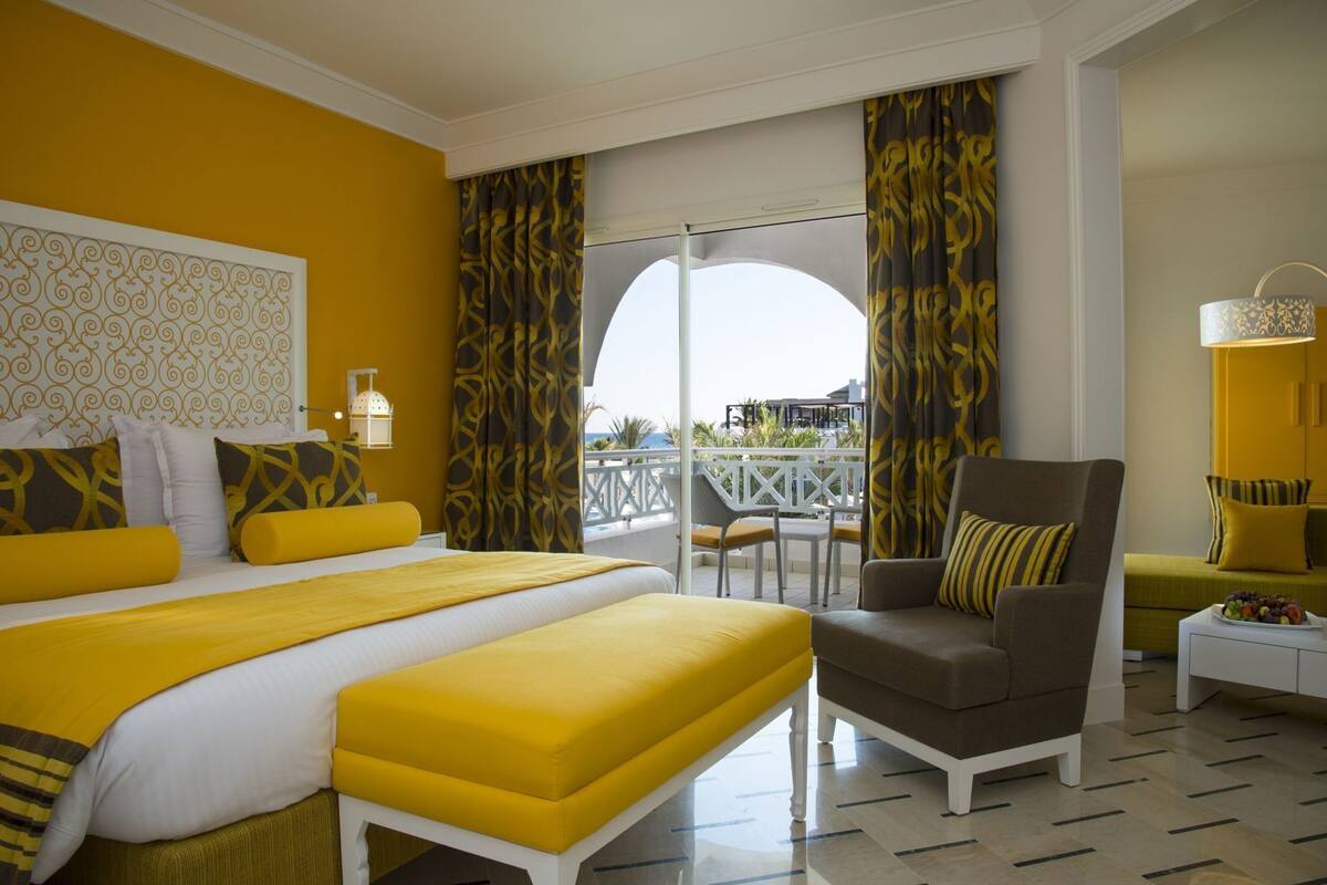 Une chambre d’hôtel avec un lit, une chaise, une petite table avec des fruits et un balcon avec vue.