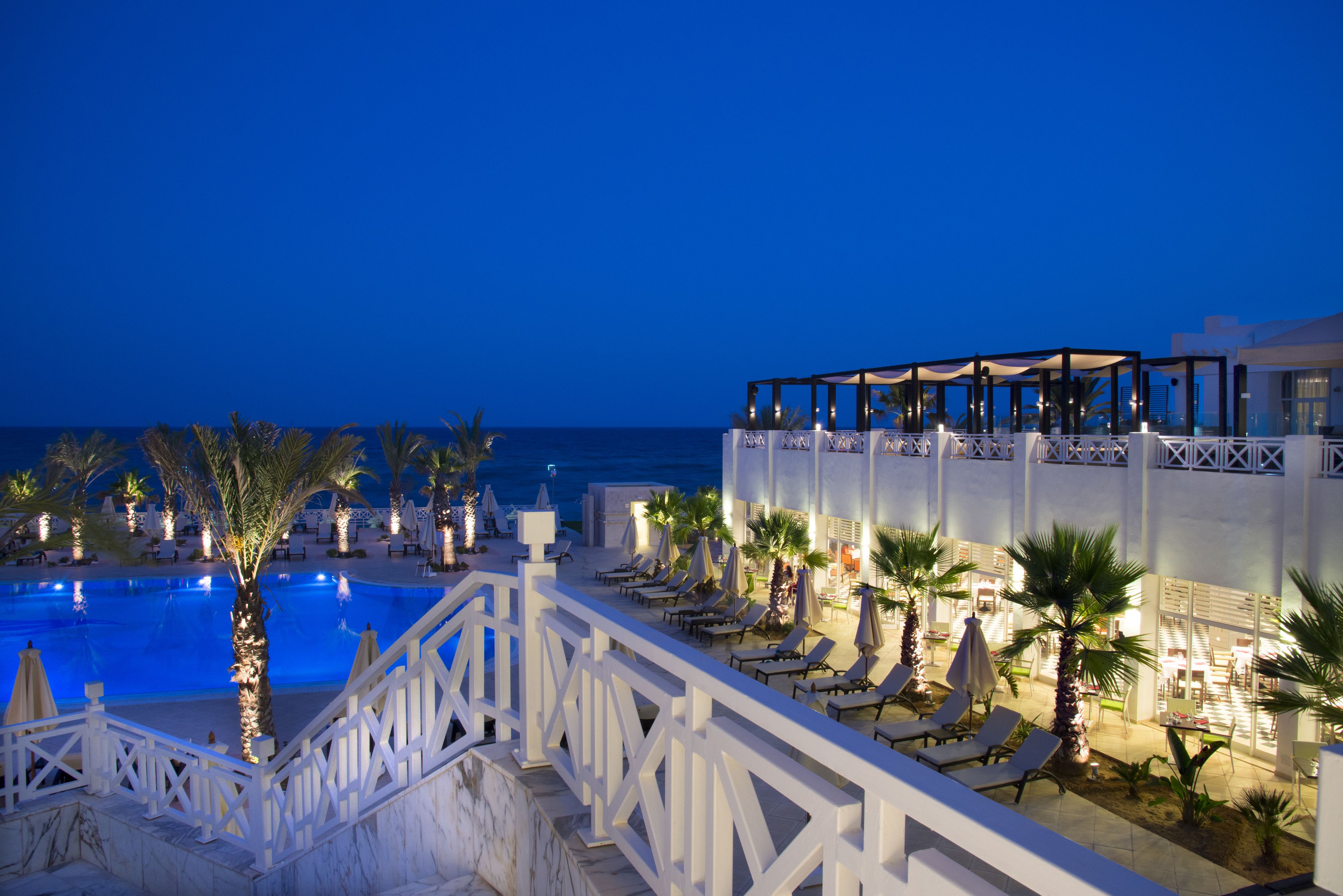 A poolside area with lounge chairs and umbrellas, a staircase leading to an upper level, and a view of the ocean at dusk.