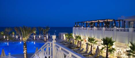 A poolside area with lounge chairs and umbrellas, a staircase leading to an upper level, and a view of the ocean at dusk.