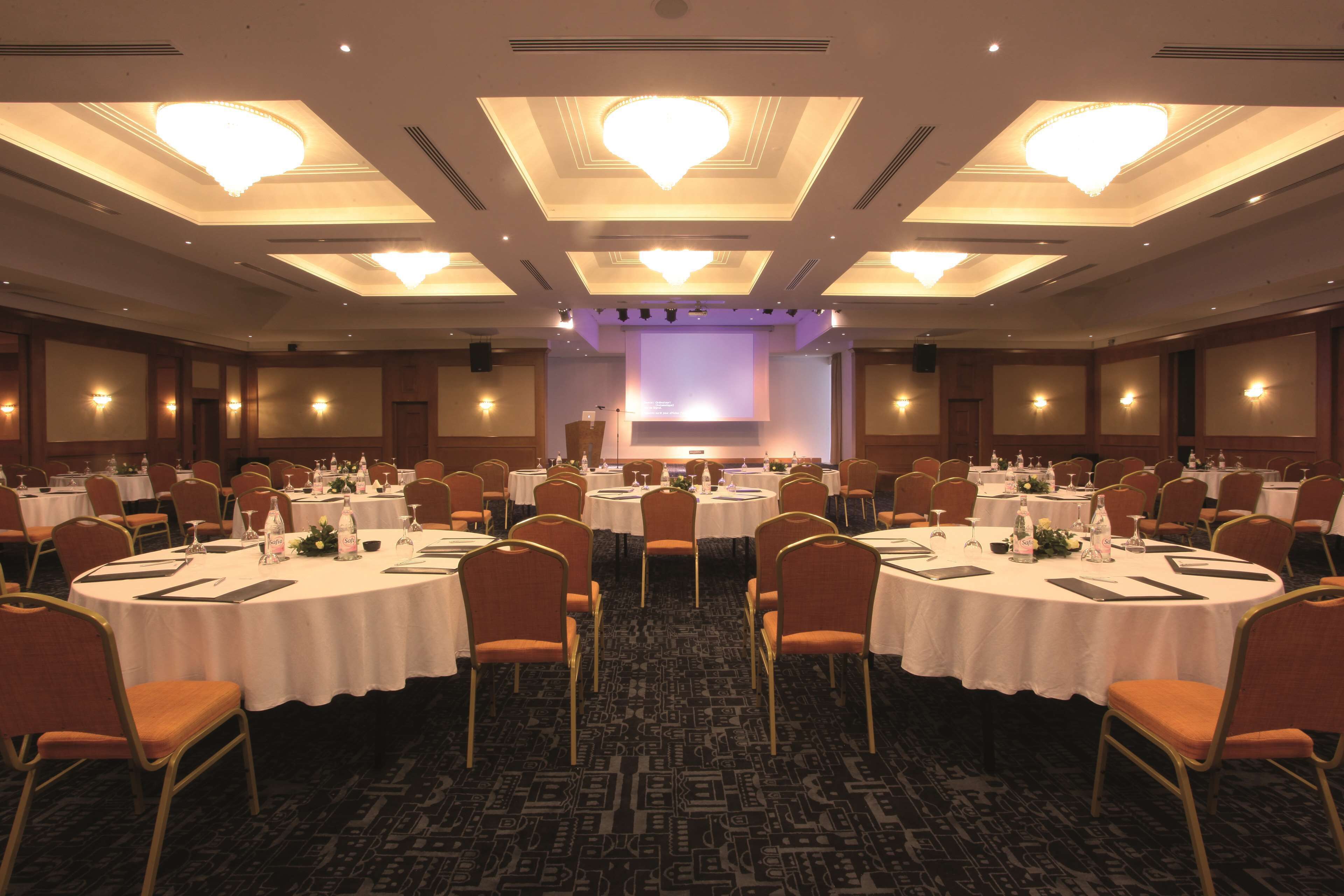 Un amplio salón de conferencias con mesas redondas, sillas y un escenario con una pantalla.