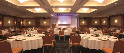 A large conference room with round tables, chairs, and a stage with a screen.