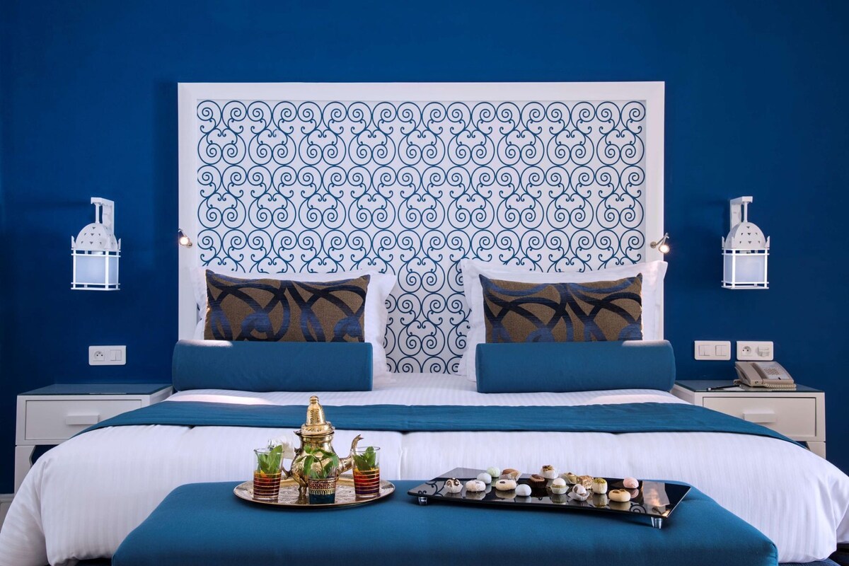 Habitación de hotel con una cama grande, dos mesitas de noche, una bandeja con bebidas y pasteles, y apliques decorativos en la pared.