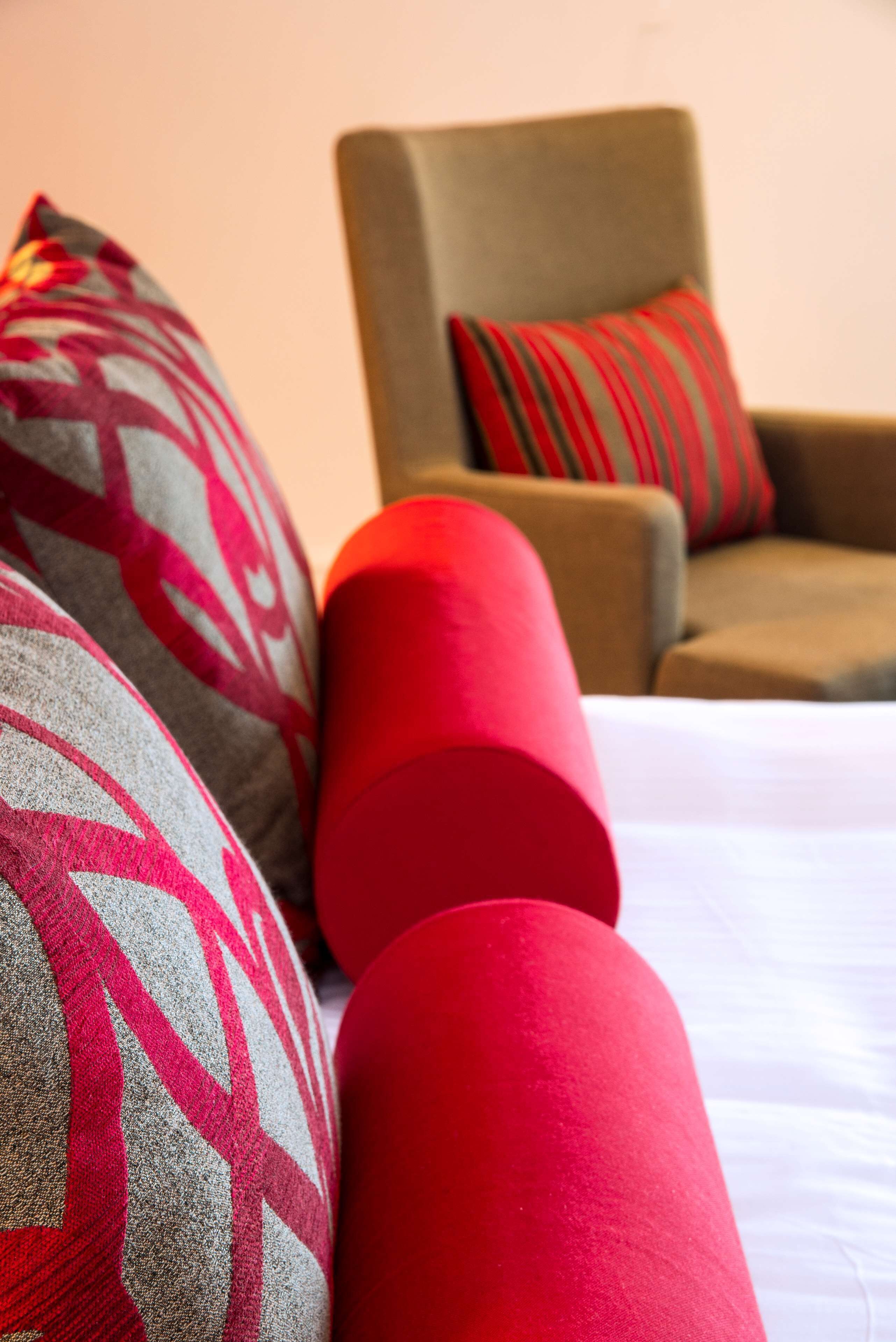Una cama con almohadas de diseño rojo y gris, un sillón marrón con un cojín a rayas, y una almohada cilíndrica roja.