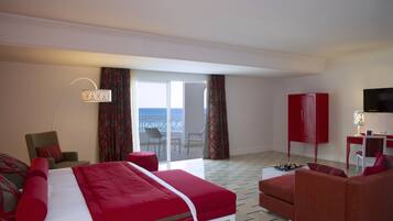Ett hotellrum med en stor säng, en soffa, ett skrivbord och en TV. Det finns en balkong med utsikt över havet.