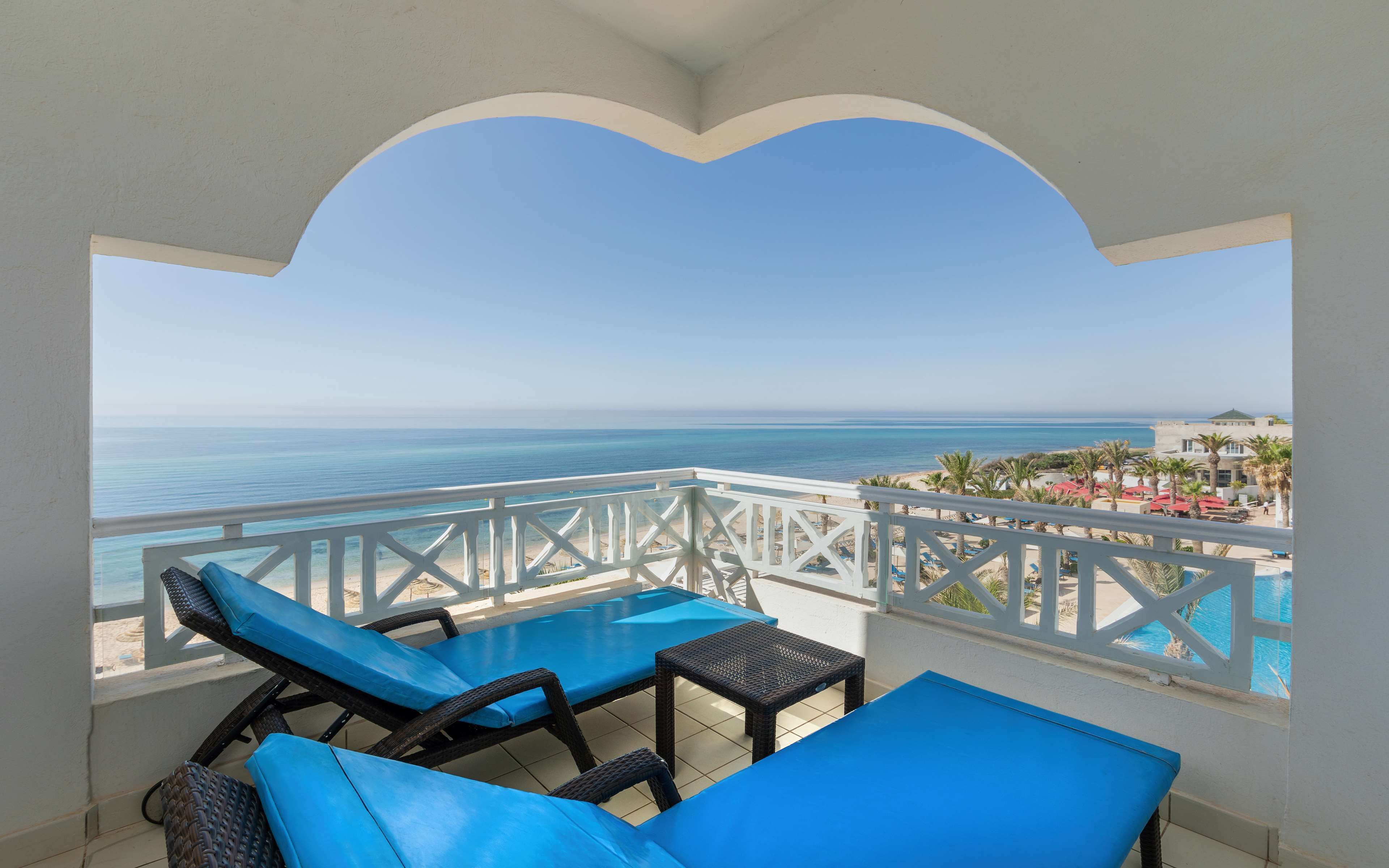 Un balcon avec des fauteuils bleus dotés de coussins et une petite table, offrant une vue sur une plage et l’océan.