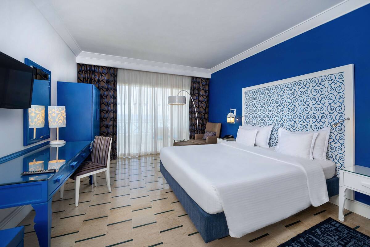 Une chambre d’hôtel avec un grand lit, un bureau avec un ordinateur, une chaise, une télévision et une fenêtre avec des rideaux.