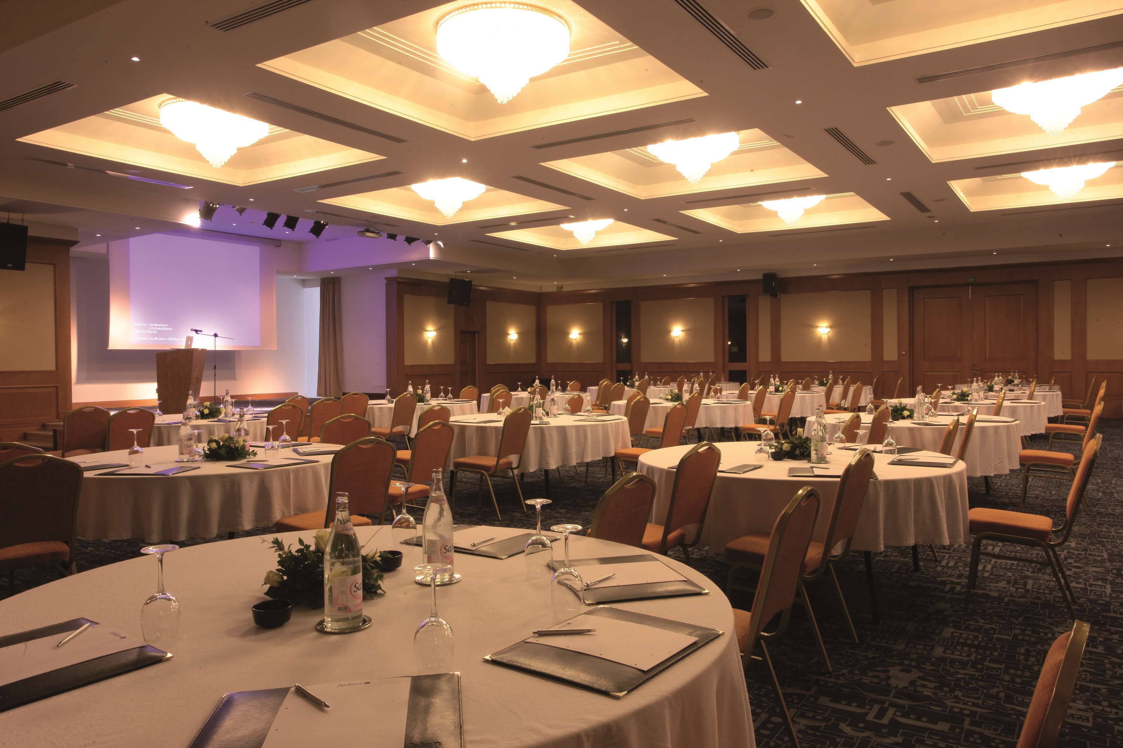 Un salón de banquetes con mesas redondas dispuestas para el servicio de comidas, equipado con sillas, vajilla y centros de mesa.