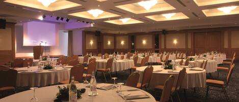 Un salón de banquetes con mesas redondas dispuestas para el servicio de comidas, equipado con sillas, vajilla y centros de mesa.
