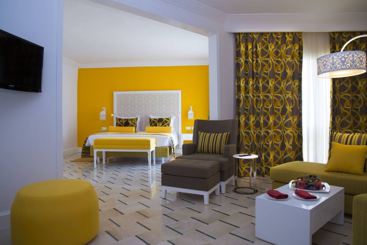 Une chambre d’hôtel avec un lit, un canapé, un pouf, une table basse avec des fruits et un téléviseur.
