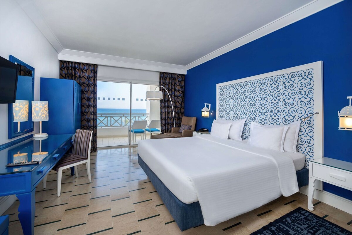 Una habitación de hotel con una cama grande, un escritorio con una silla, un televisor y un balcón con vistas al mar.