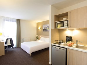 Une chambre d’hôtel équipée d’un lit, d’un bureau, d’une chaise, d’une kitchenette et d’un petit réfrigérateur.