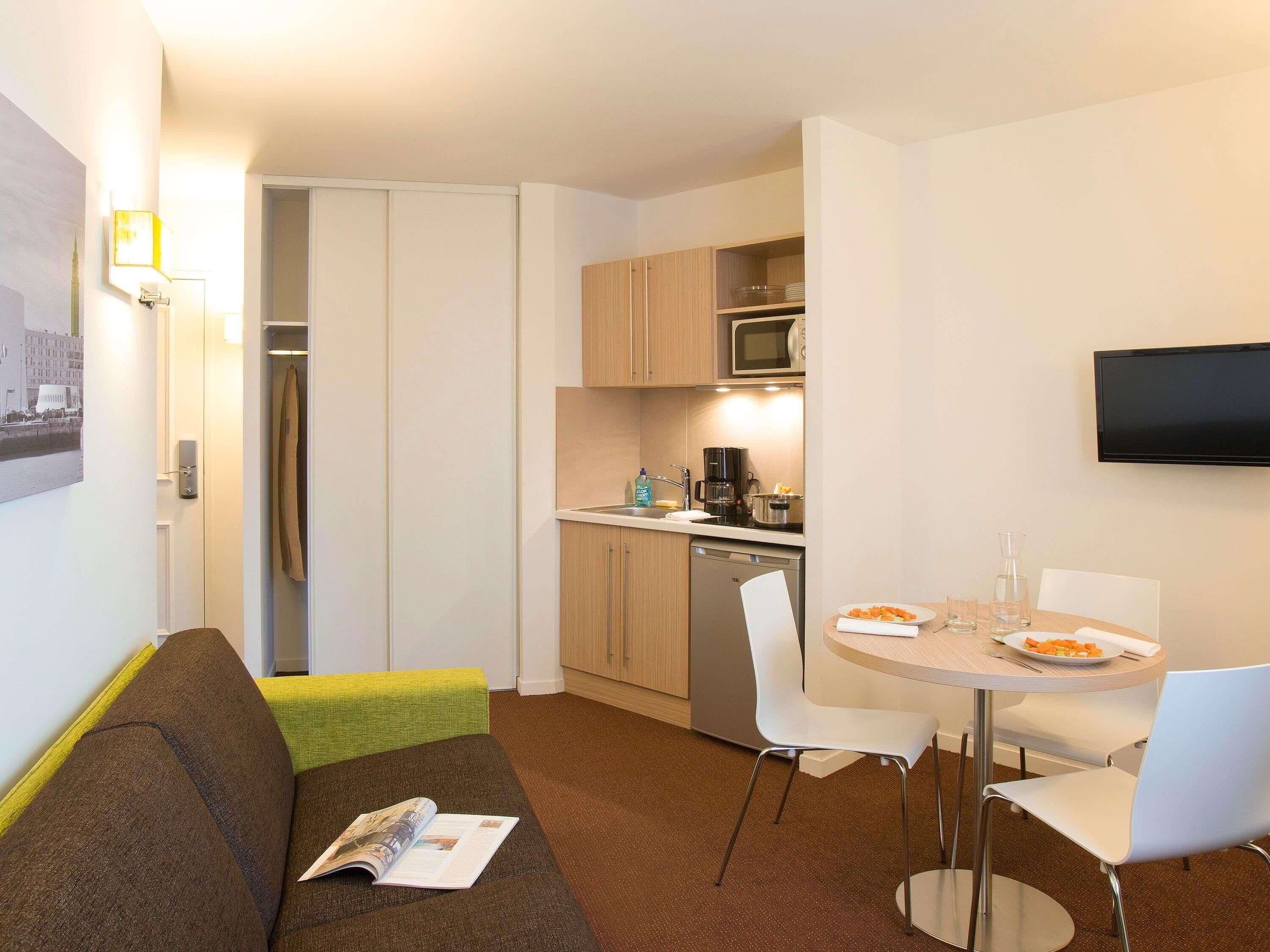 A compact hotel room with a kitchenette, a dining area with a table and chairs, a sofa with a magazine, and a flat-screen TV mounted on the wall.