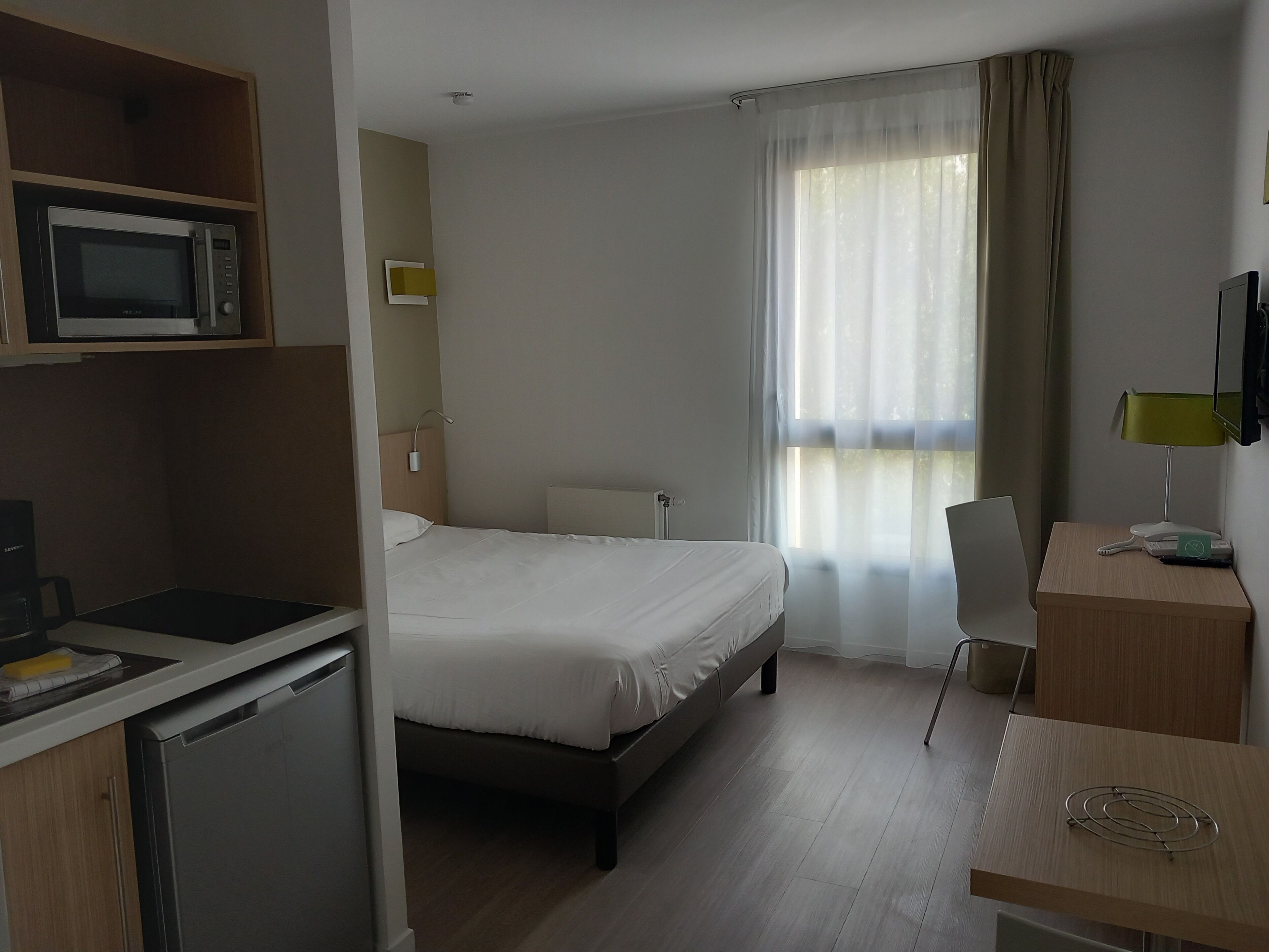 A hotel room with a bed, a desk, a chair, a microwave, a television, and a window with curtains.