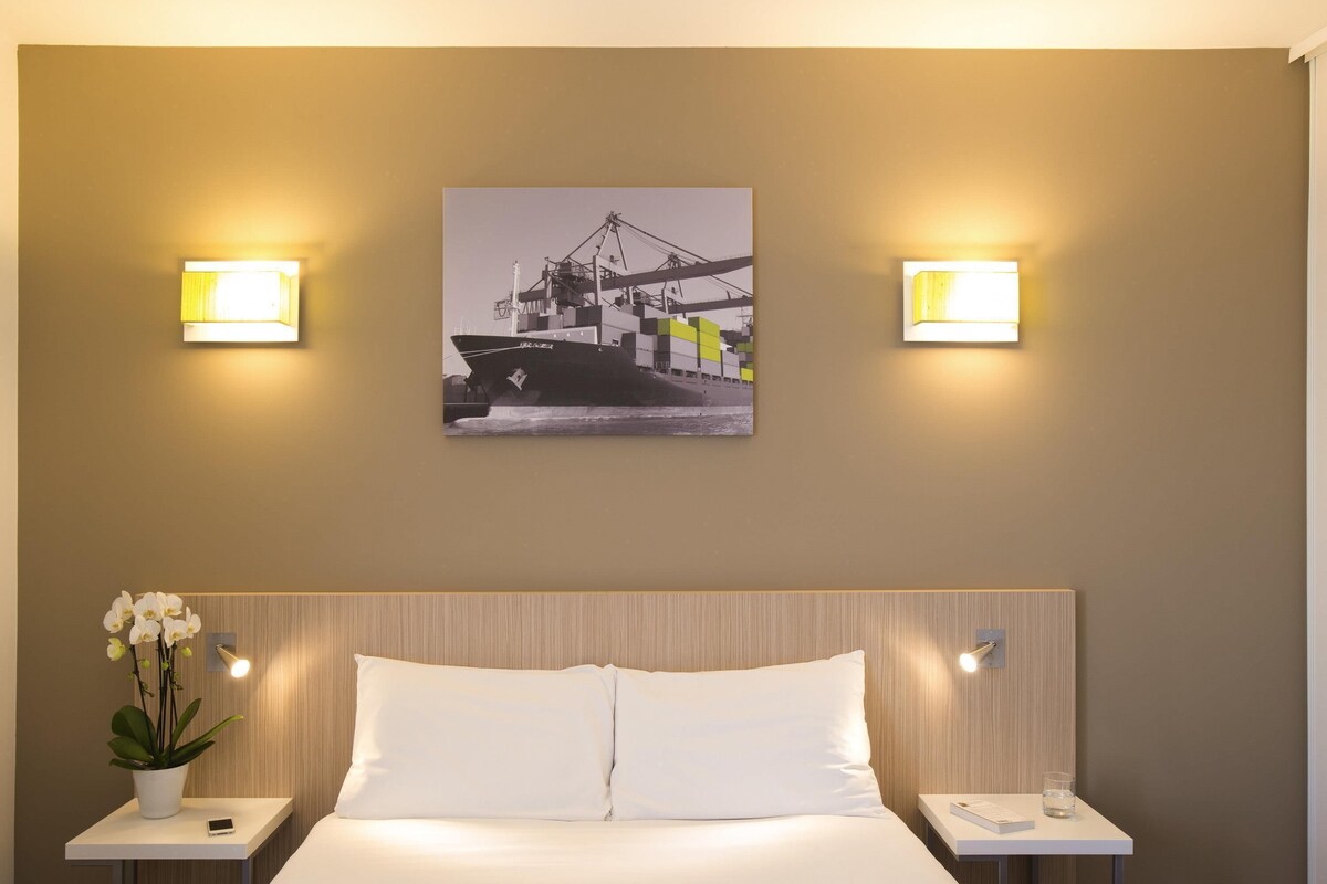 A hotel room with a bed, two bedside tables, a nightstand, a wall-mounted picture, and two wall-mounted lights.