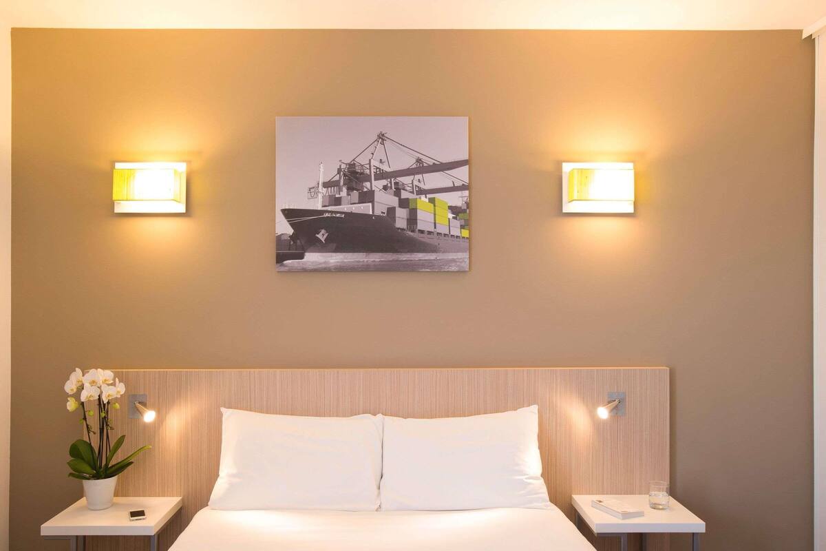 A hotel room with a bed, two bedside tables, a nightstand, a wall-mounted picture, and two wall-mounted lights.