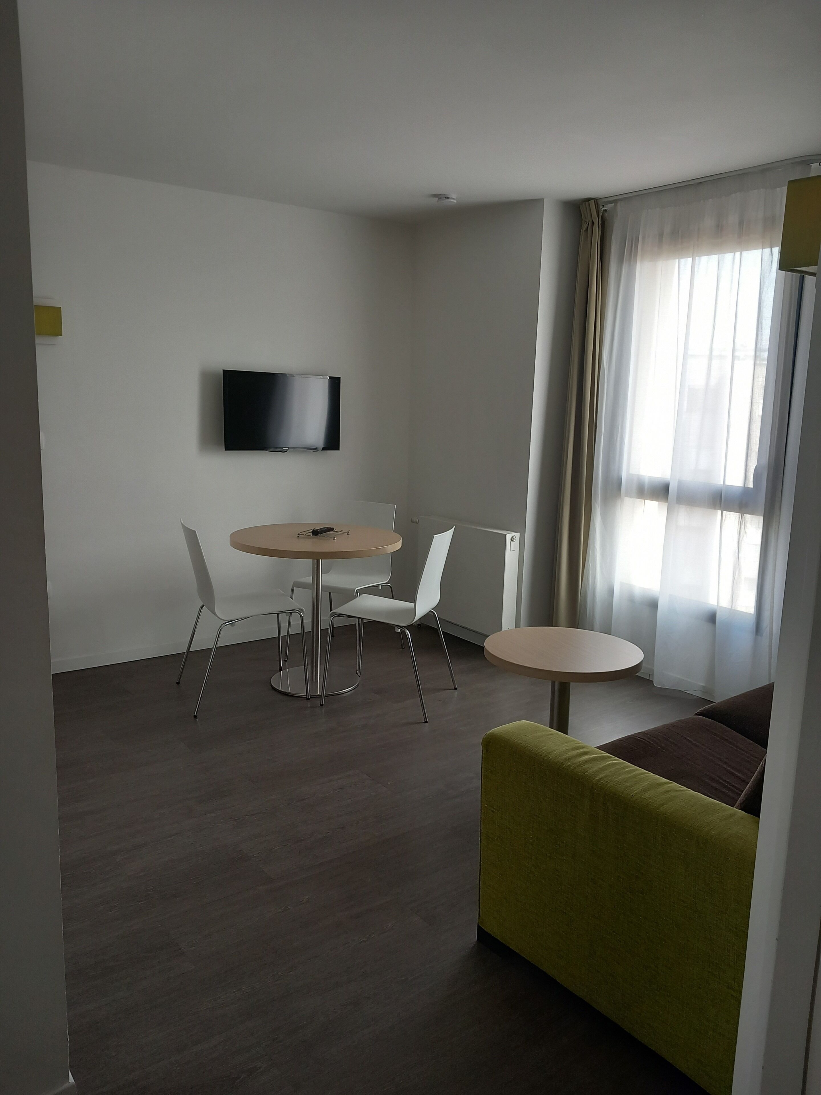 Uma sala de estar moderna com televisão de tela plana na parede, uma mesa de jantar redonda com cadeiras, um sofá e uma janela com cortinas leves.