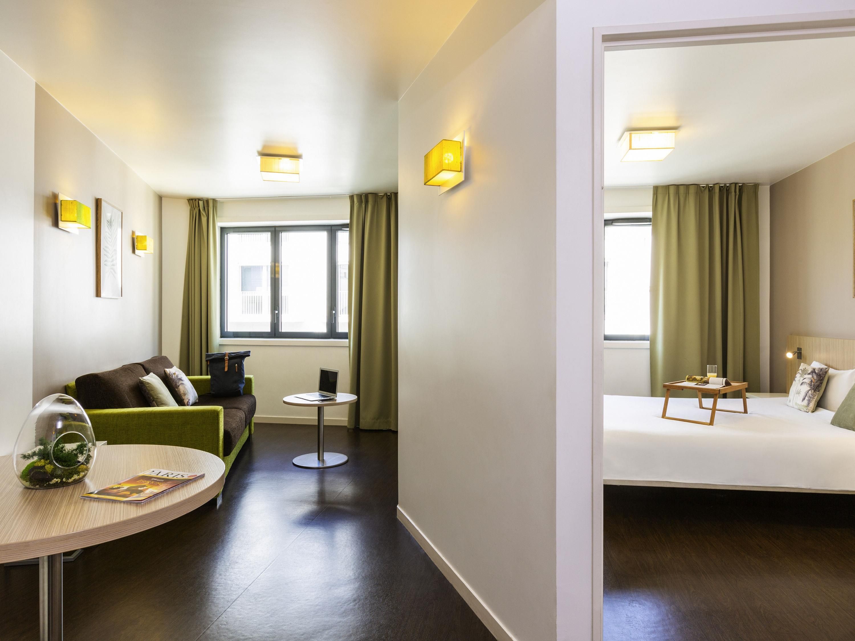 Une chambre d’hôtel avec un canapé vert, une petite table ronde sur laquelle se trouve un ordinateur portable, un lit avec une table de chevet et une fenêtre avec des rideaux.