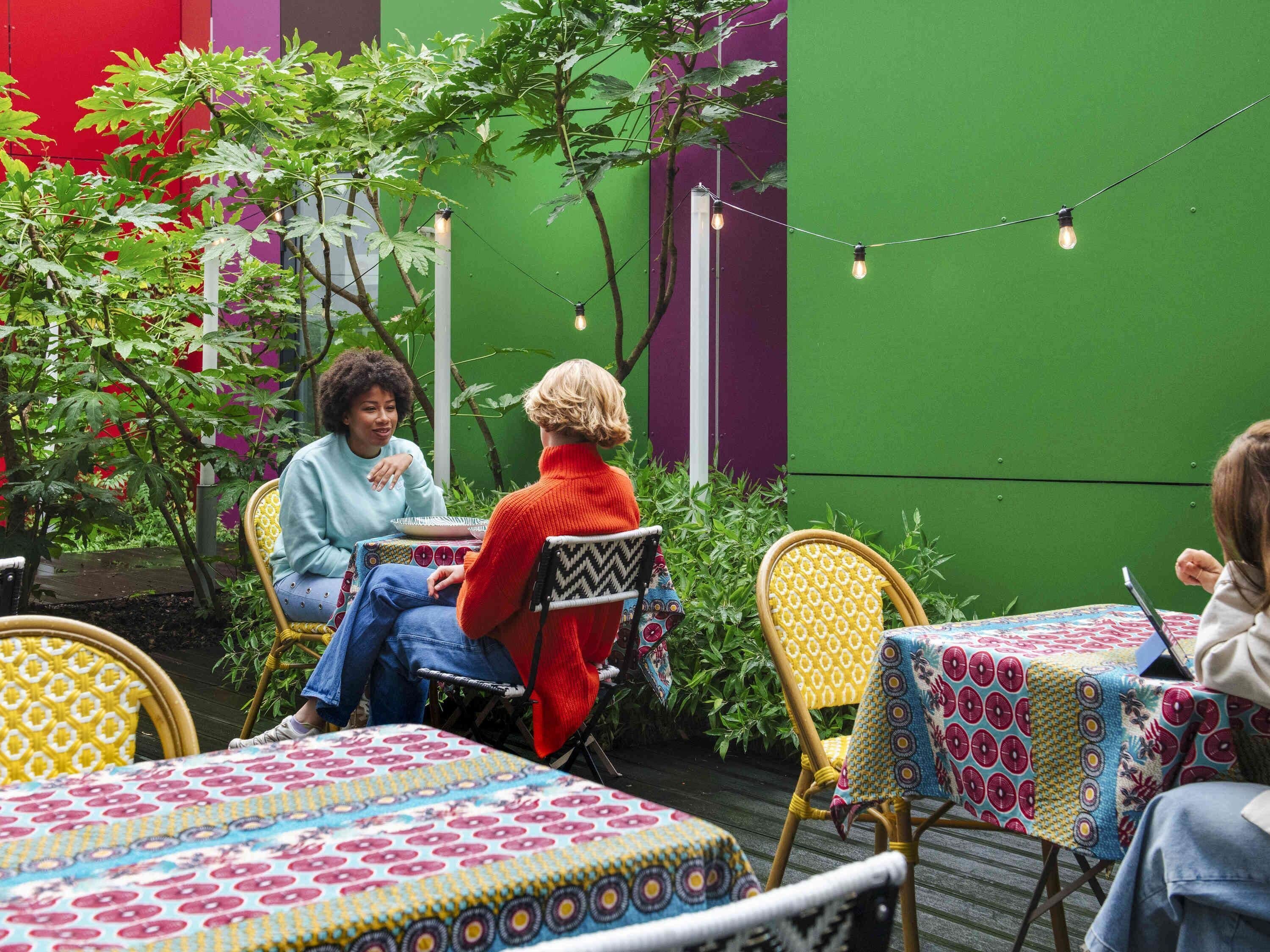 Un espacio al aire libre con mesas y sillas, rodeado de follaje y una pared verde.