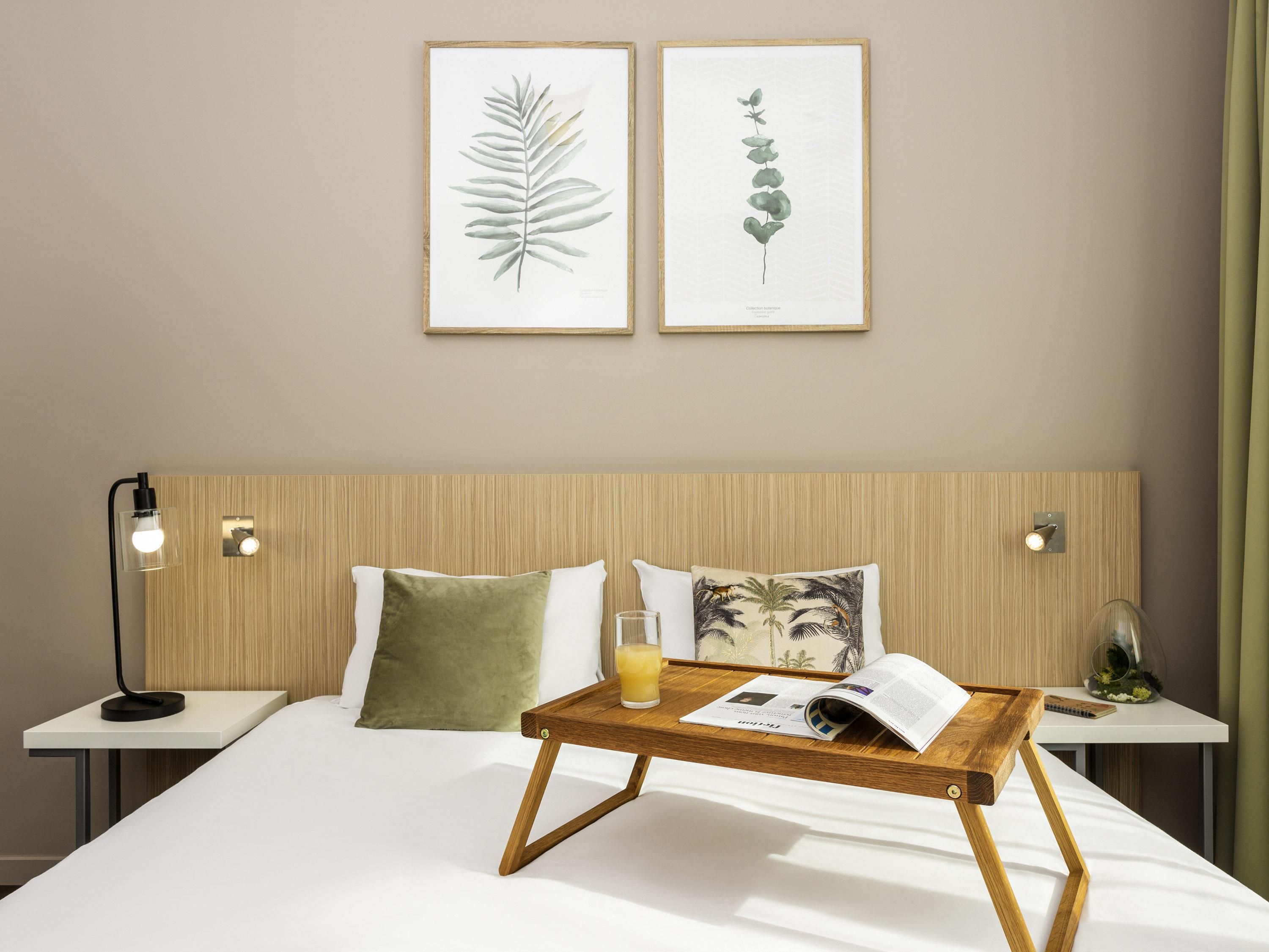 A hotel room with a wooden headboard, two bedside tables, a bed with white bedding, a wooden side table with a glass of juice, and two framed botanical prints on the wall.