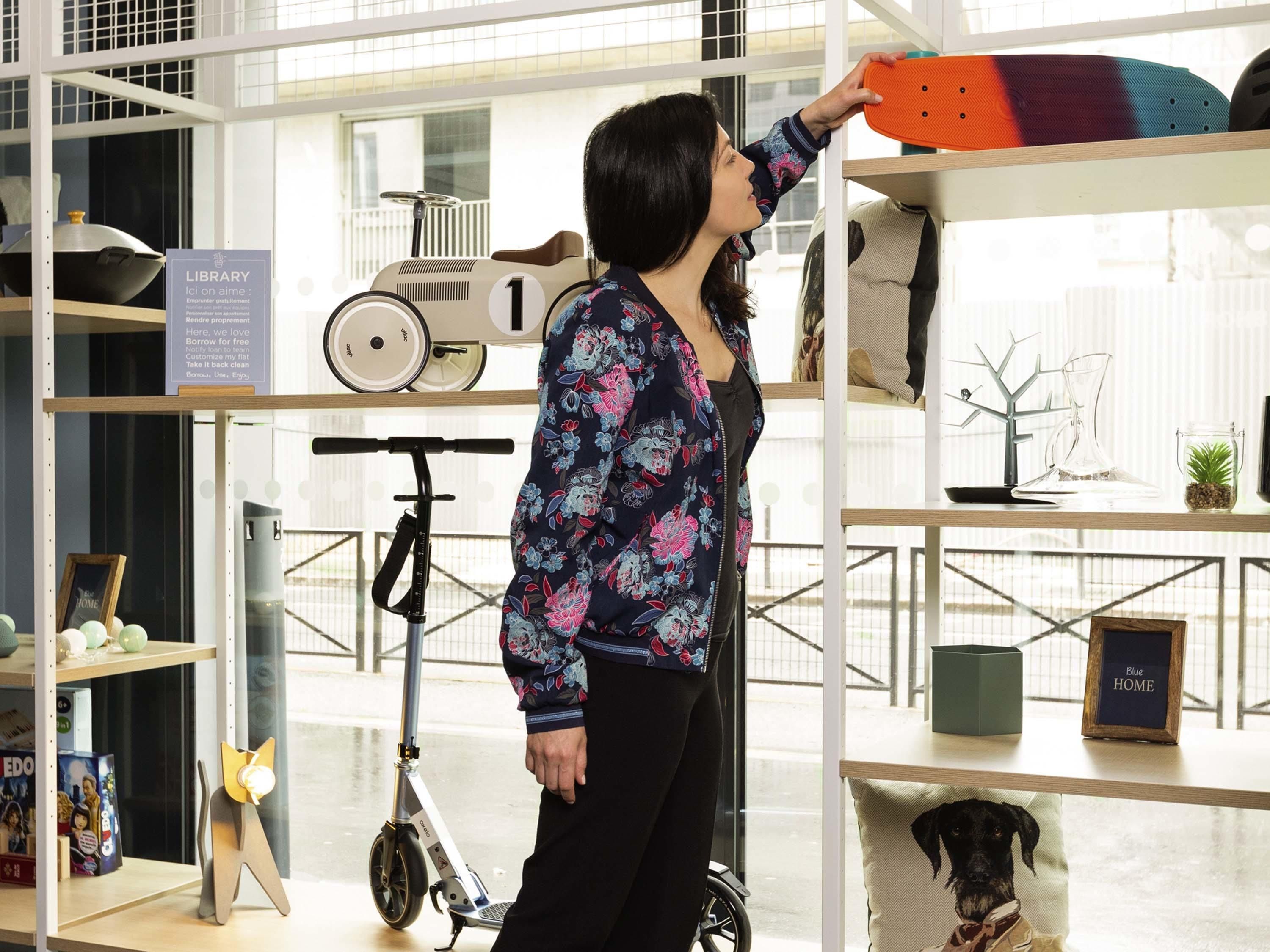 Eine Person mit Blumenbluse blättert an einem Bücherregal mit Büchern und einem Rollstuhl.