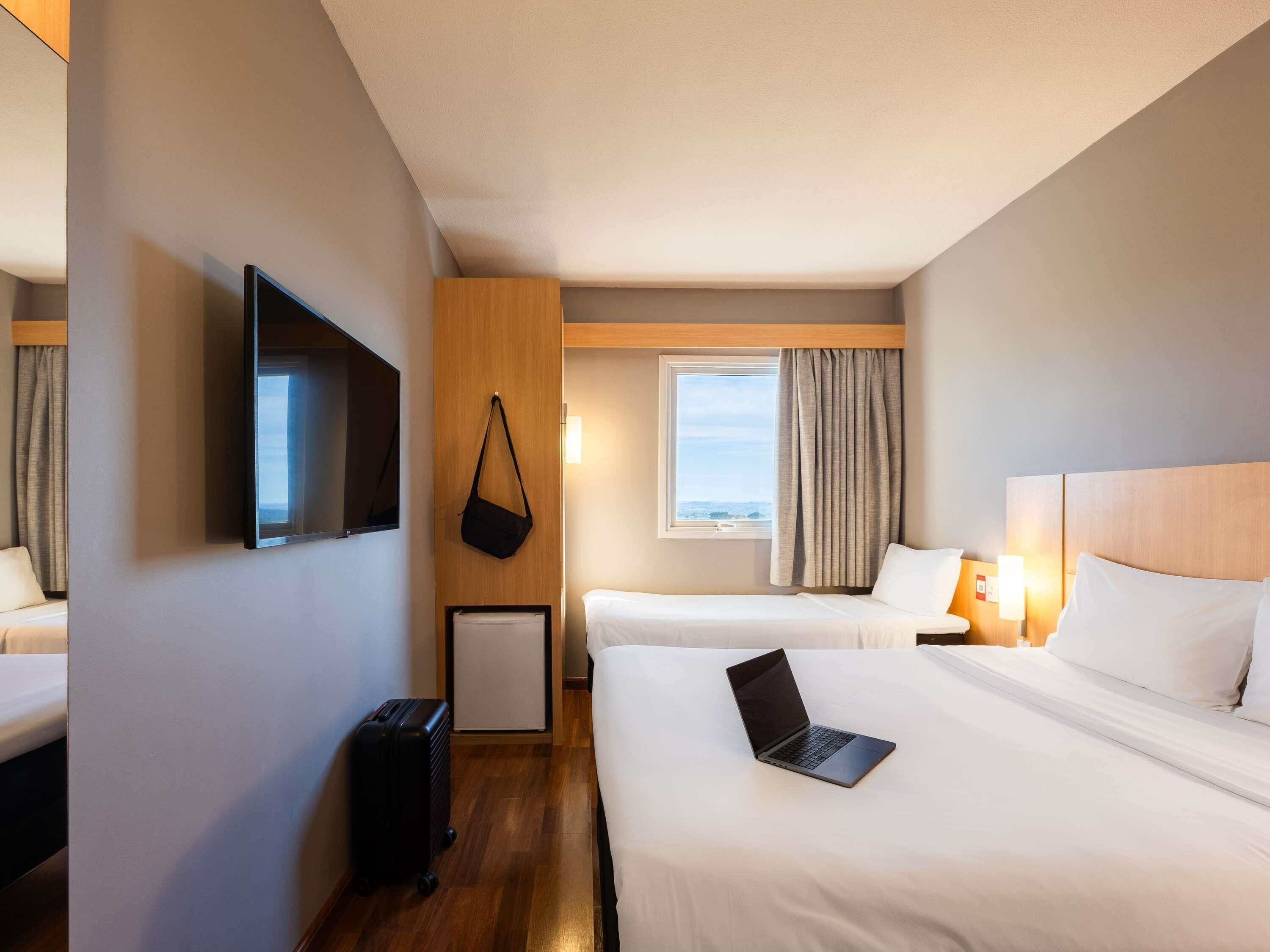 A hotel room with two beds, a TV, a window with curtains, a desk with a lamp, and a suitcase.