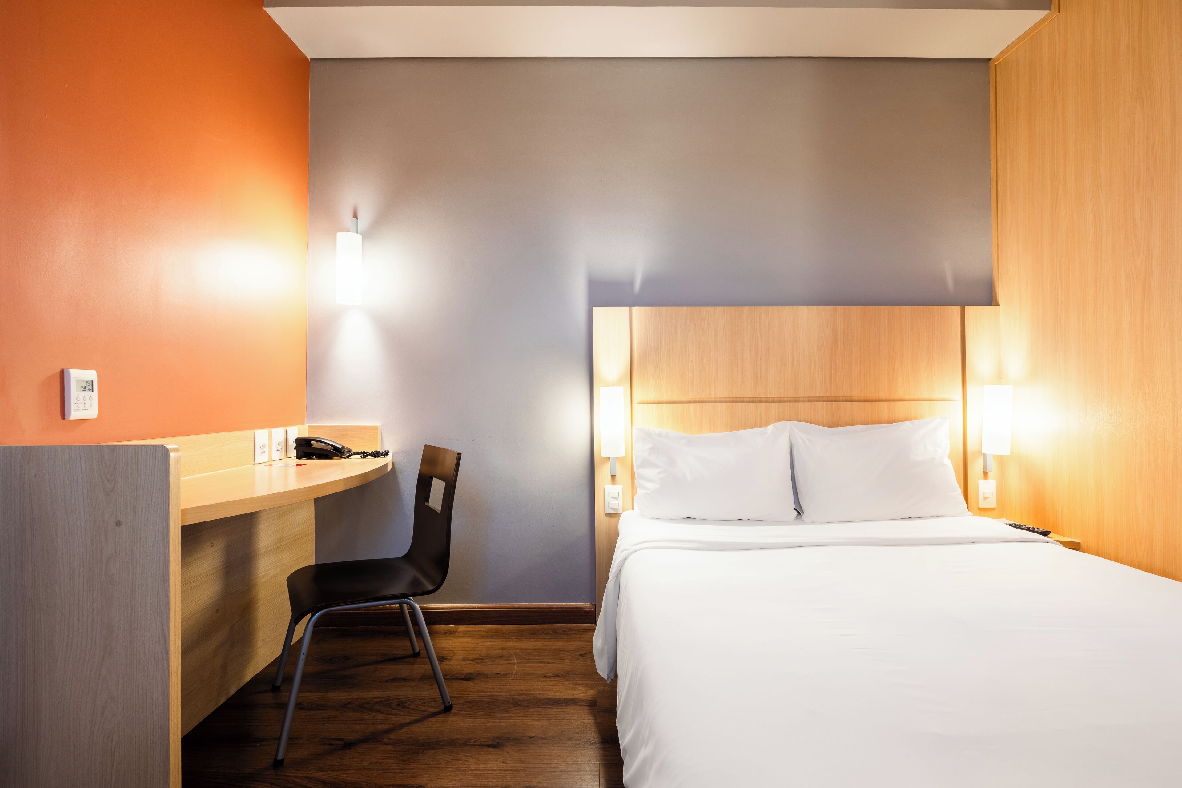 Habitación de hotel con un escritorio de madera, una silla, una cama con ropa blanca y dos lámparas fijadas a la pared.
