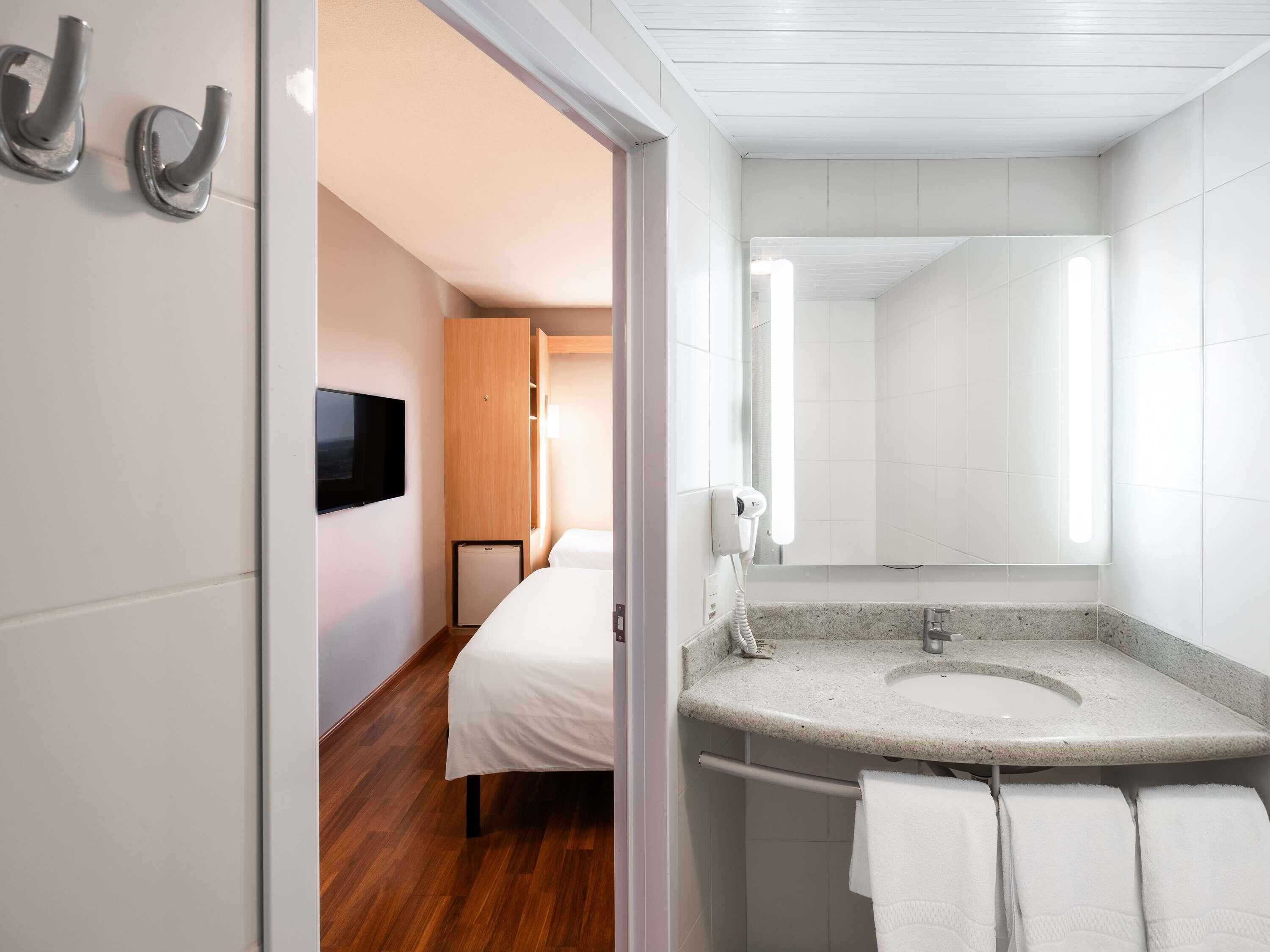 A modern hotel room with a bed, a small TV, a bathroom with a sink and mirror, and a wooden wardrobe.