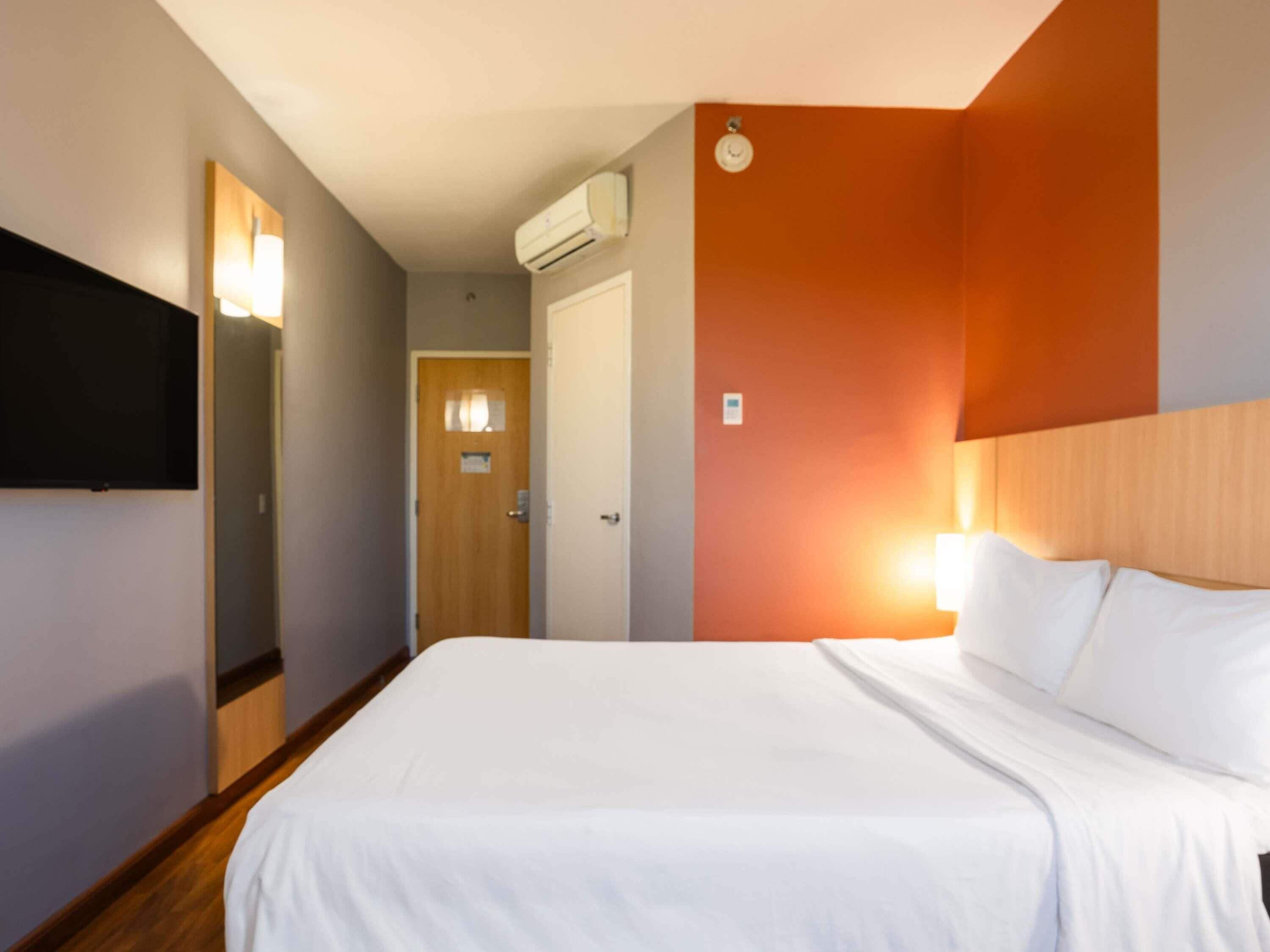 A hotel room with a bed, a television, an air conditioning unit, and a door leading to another room.