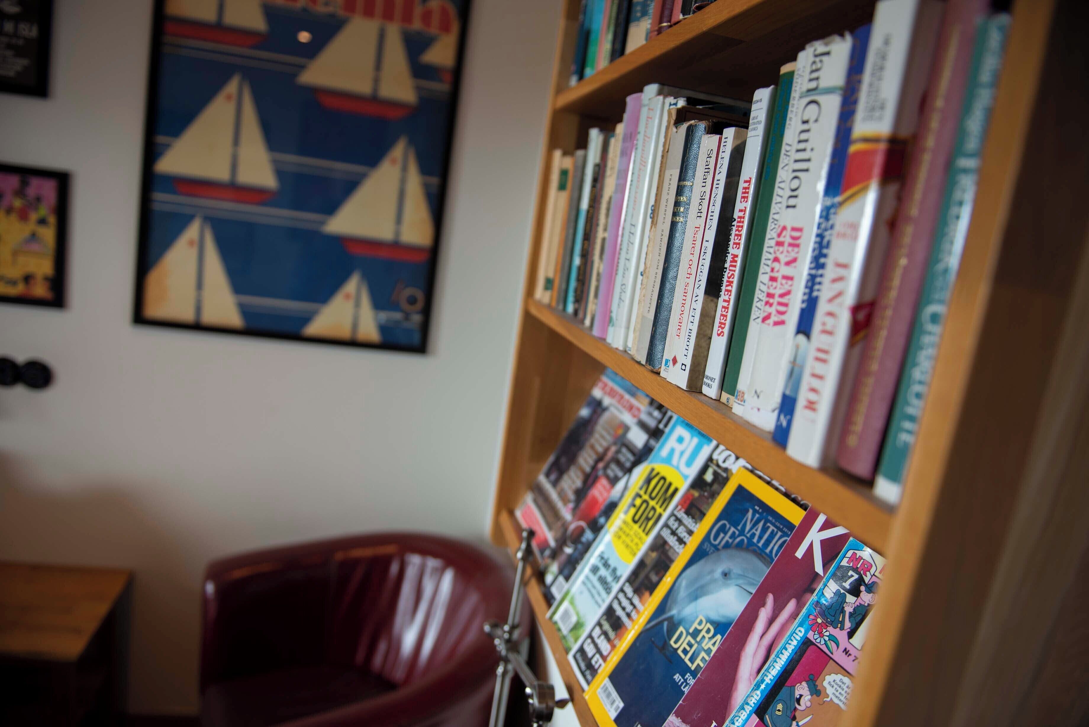 A bookshelf filled with books and magazines, a red chair, and a sailboat painting on the wall.