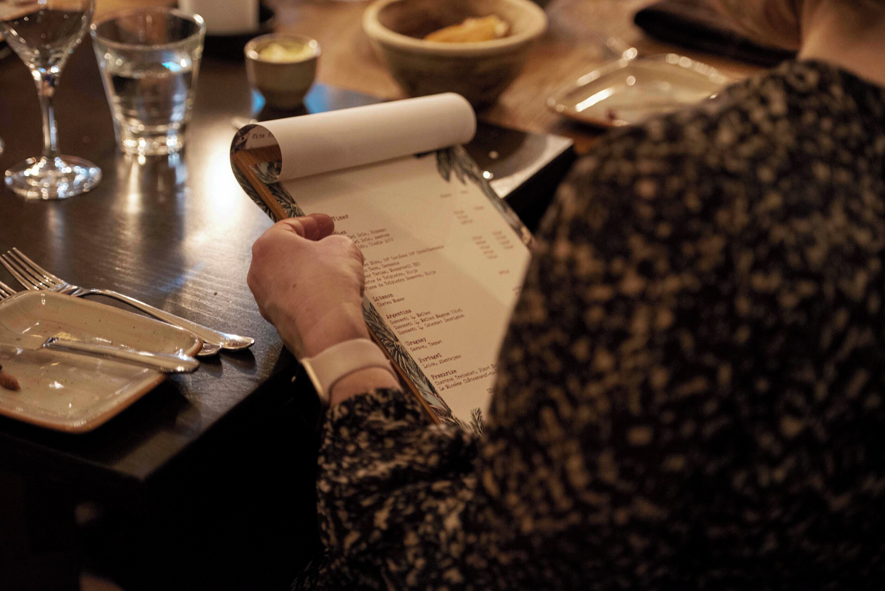 A person holding a menu at a table with cutlery and a glass of water.