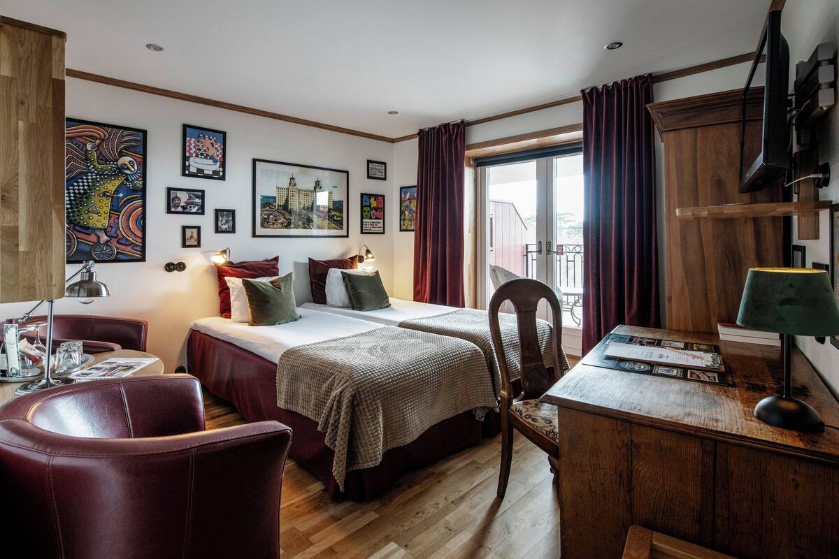 A hotel room with two beds, a desk, a chair, a wooden floor, and artwork on the walls.