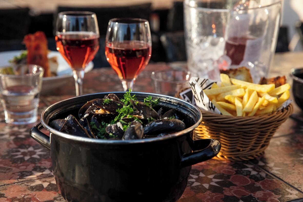 Uma mesa de jantar com um pote preto de mexilhões, dois copos de vinho tinto, uma cesta de batatas fritas e um copo de água.