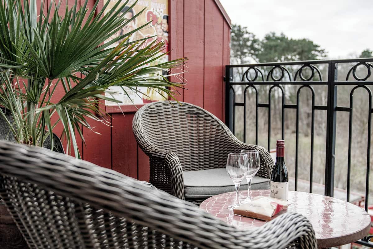 A wicker table with two wine glasses and a bottle on a deck with a red building in the background.