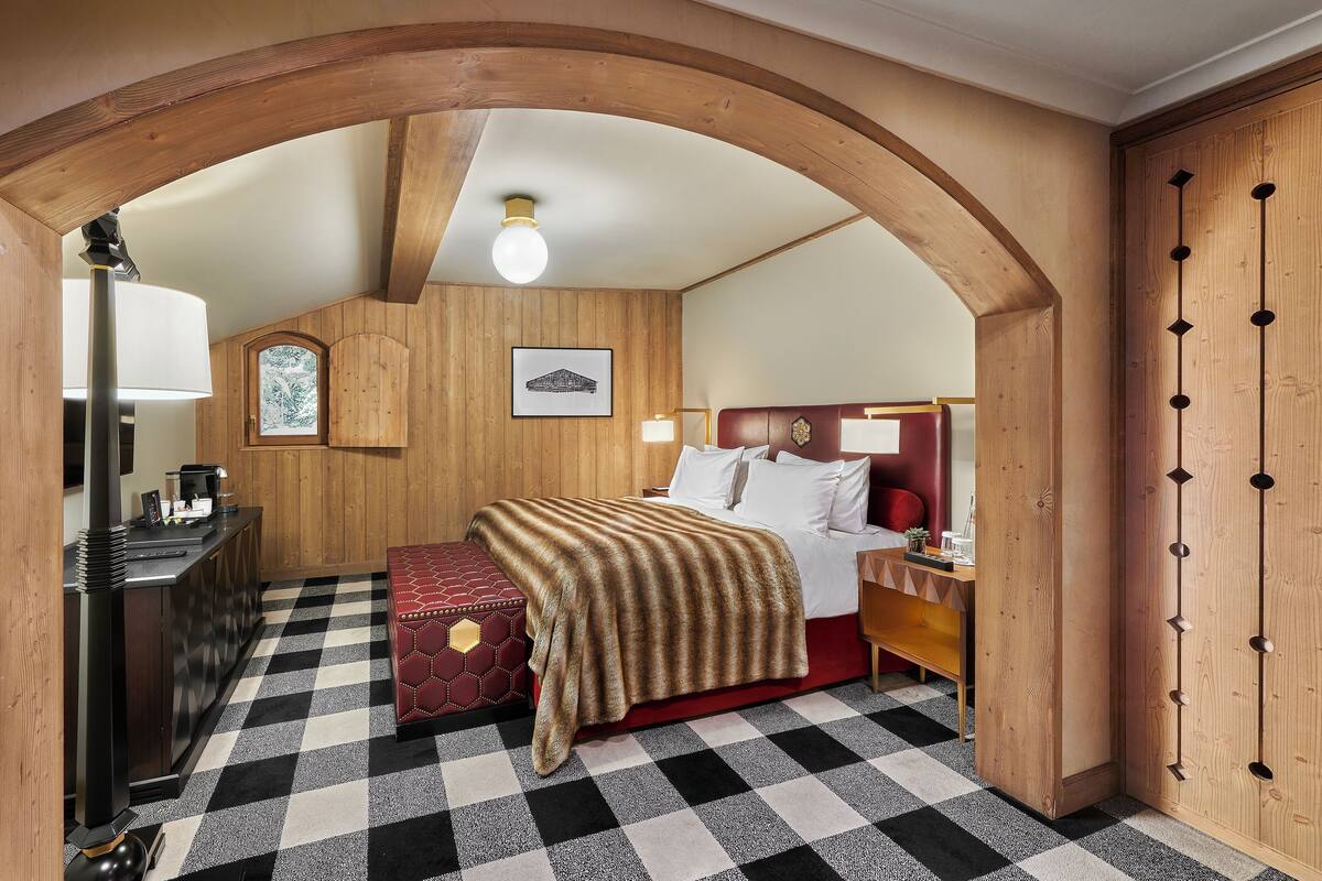 Ein Hotelzimmer mit Schachbrettmuster-Boden, Holzwänden, einem Bett mit rotem Kopfteil, einer roten Bank, einem Holzschrank und einem Holzbogen über der Tür.