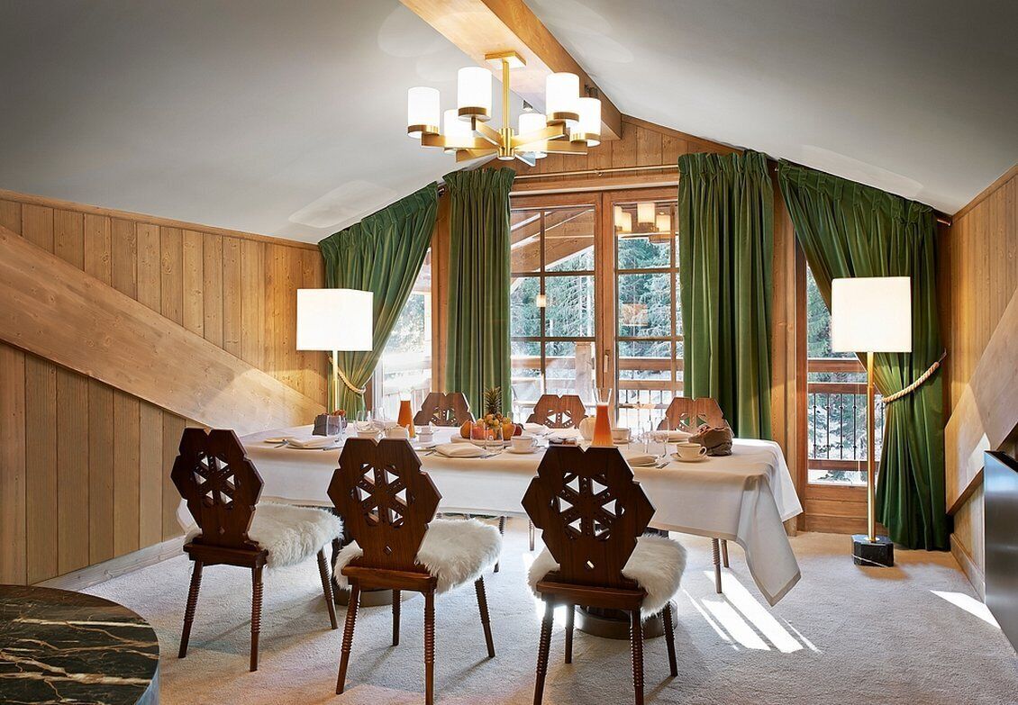 A dining room with a high ceiling, wooden walls, and a large window with green curtains. There are four wooden chairs with white fur throws and a table set for four.