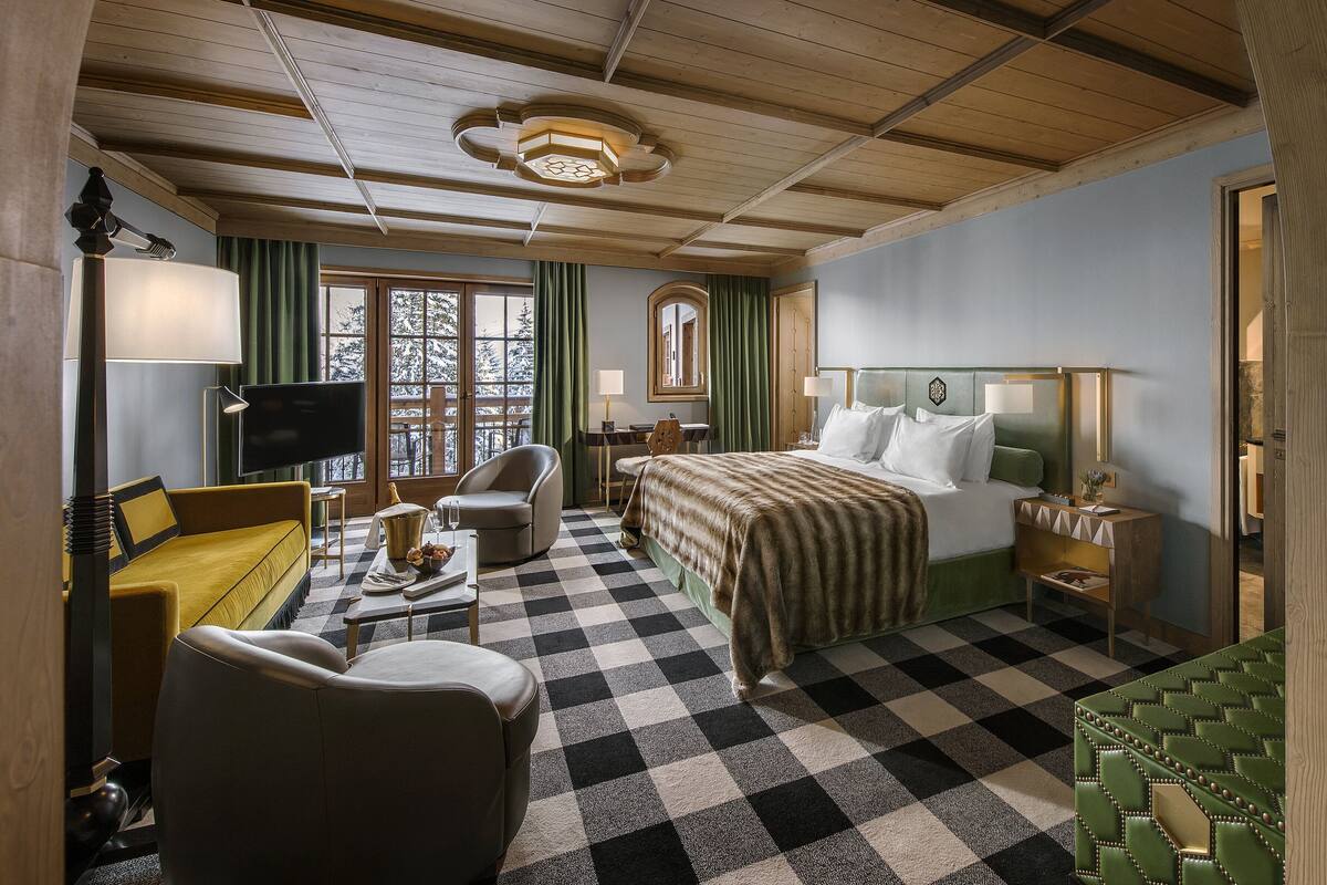 Ein Hotelzimmer mit Schachbrettmuster-Boden, einem Bett mit grünem Kopfteil, einem Schreibtisch mit Lampe, einem Sessel und einem Fernseher.