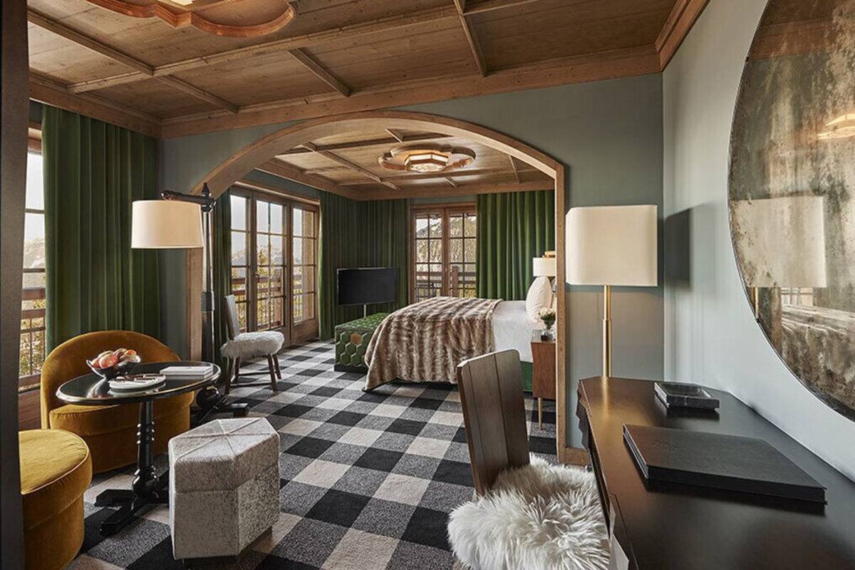 Ein Hotelzimmer mit Schachbrettmuster-Boden, einem Bett mit gestreifter Decke, einem Schreibtisch mit Lampe, einem Sessel und einem großen Fenster.
