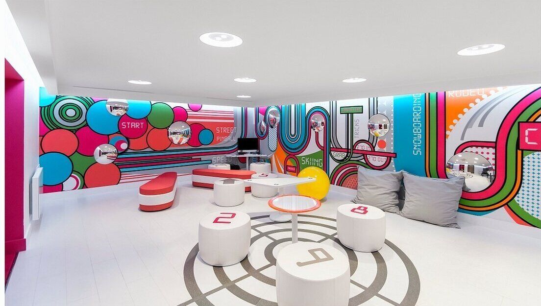 A brightly colored room with a playful design, featuring a large wall mural, multiple seating areas, and a central table.