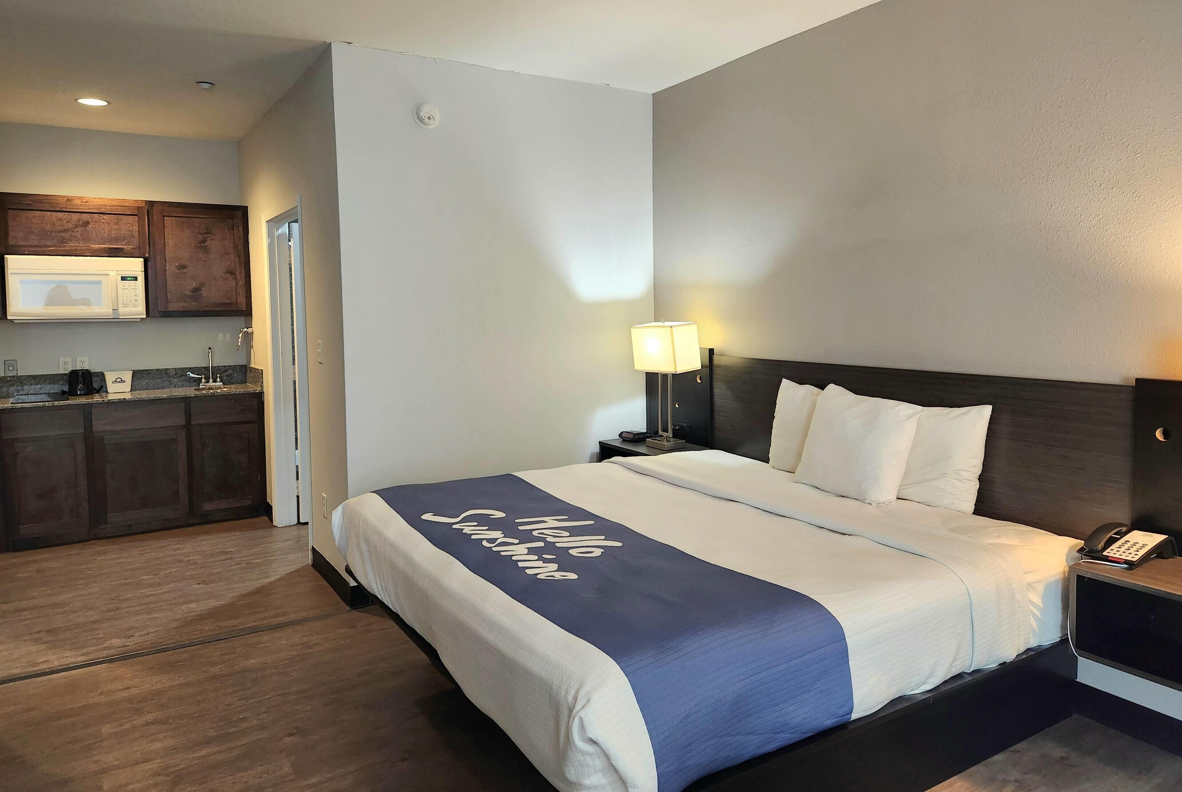 Quarto de hotel com cama, mesa de cabeceira, luminária e vista para a cozinha.