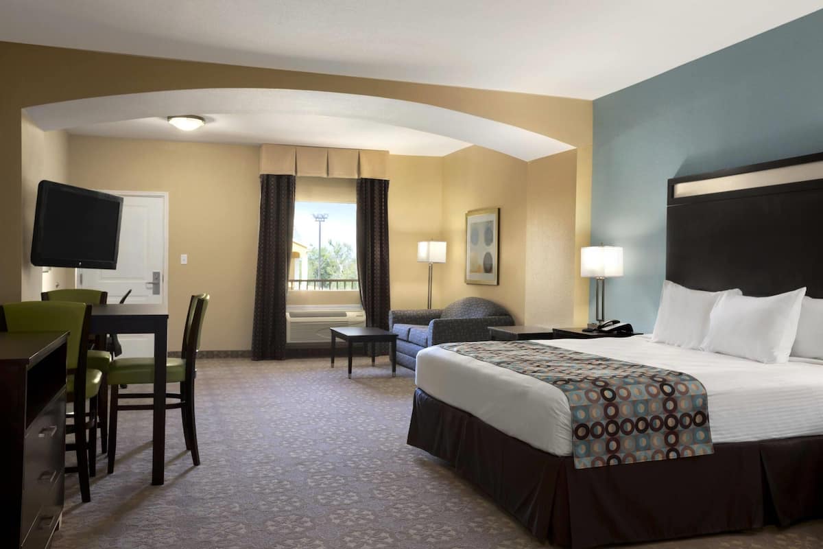 Quarto de hotel com cama, escrivaninha, cadeiras, televisão e janela com cortinas.
