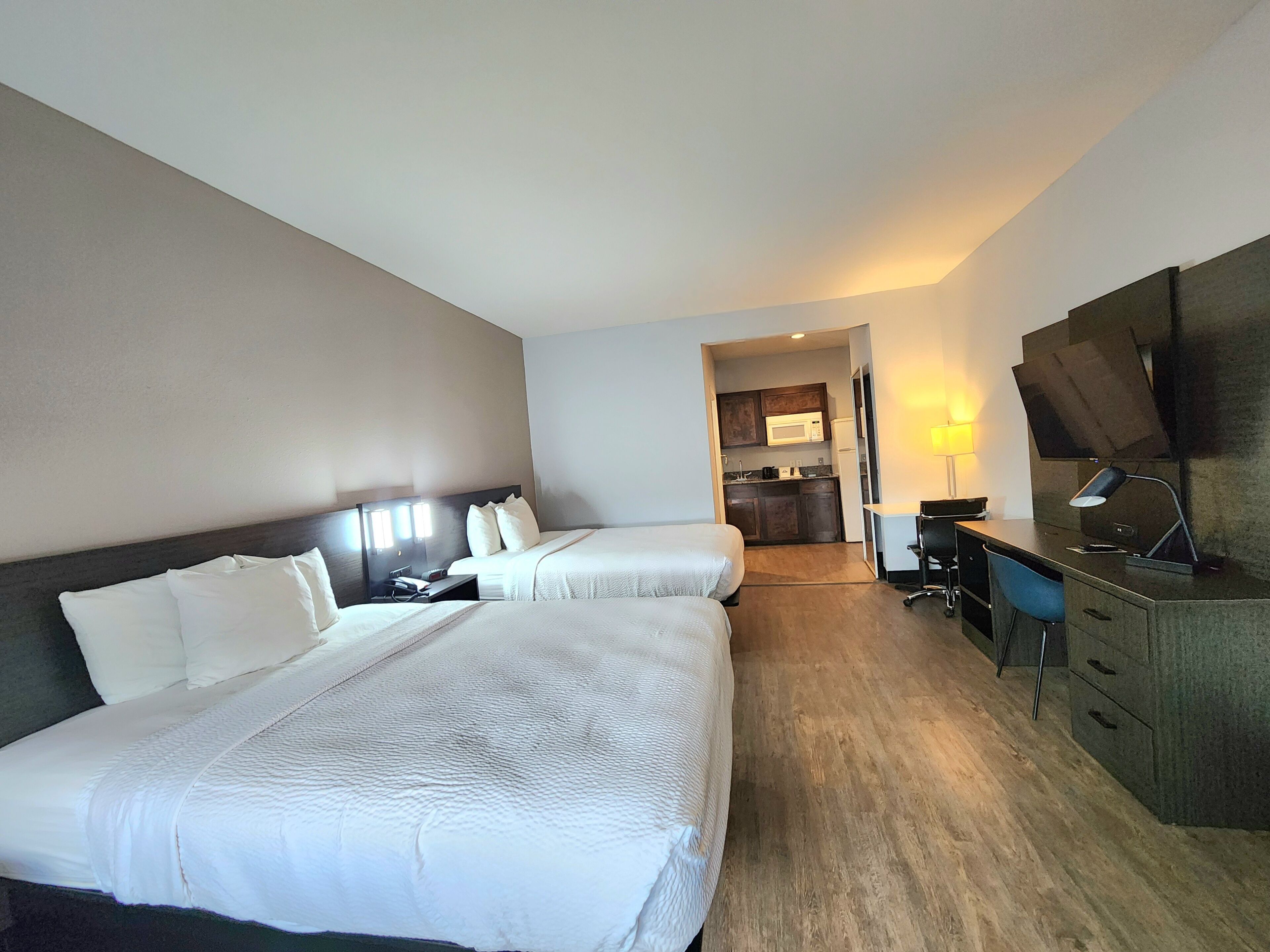 A hotel room with two beds, a desk, a chair, a TV, and a kitchenette.