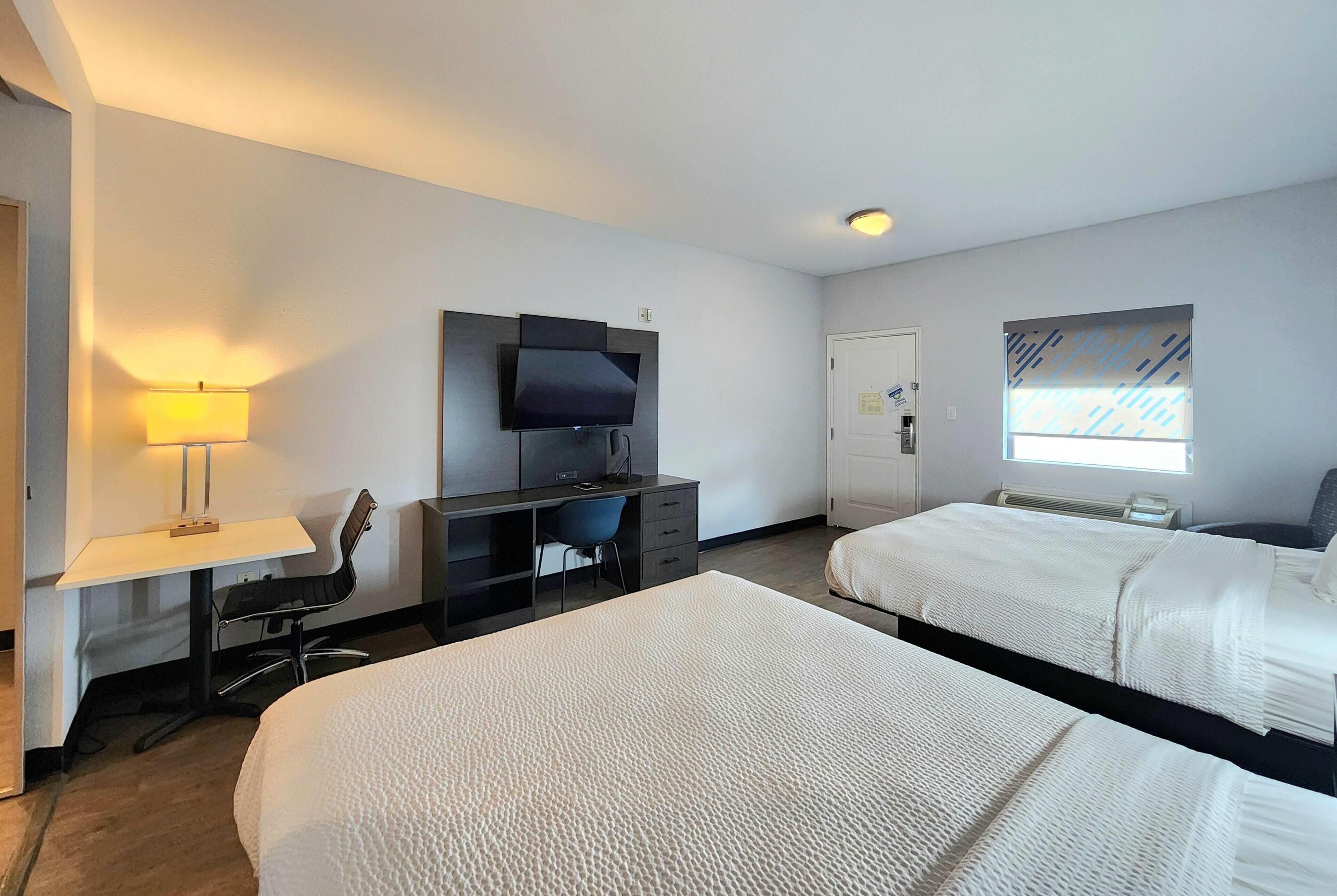 Une chambre d’hôtel avec deux lits, un bureau avec une chaise, une télévision, une lampe et une fenêtre avec des stores.
