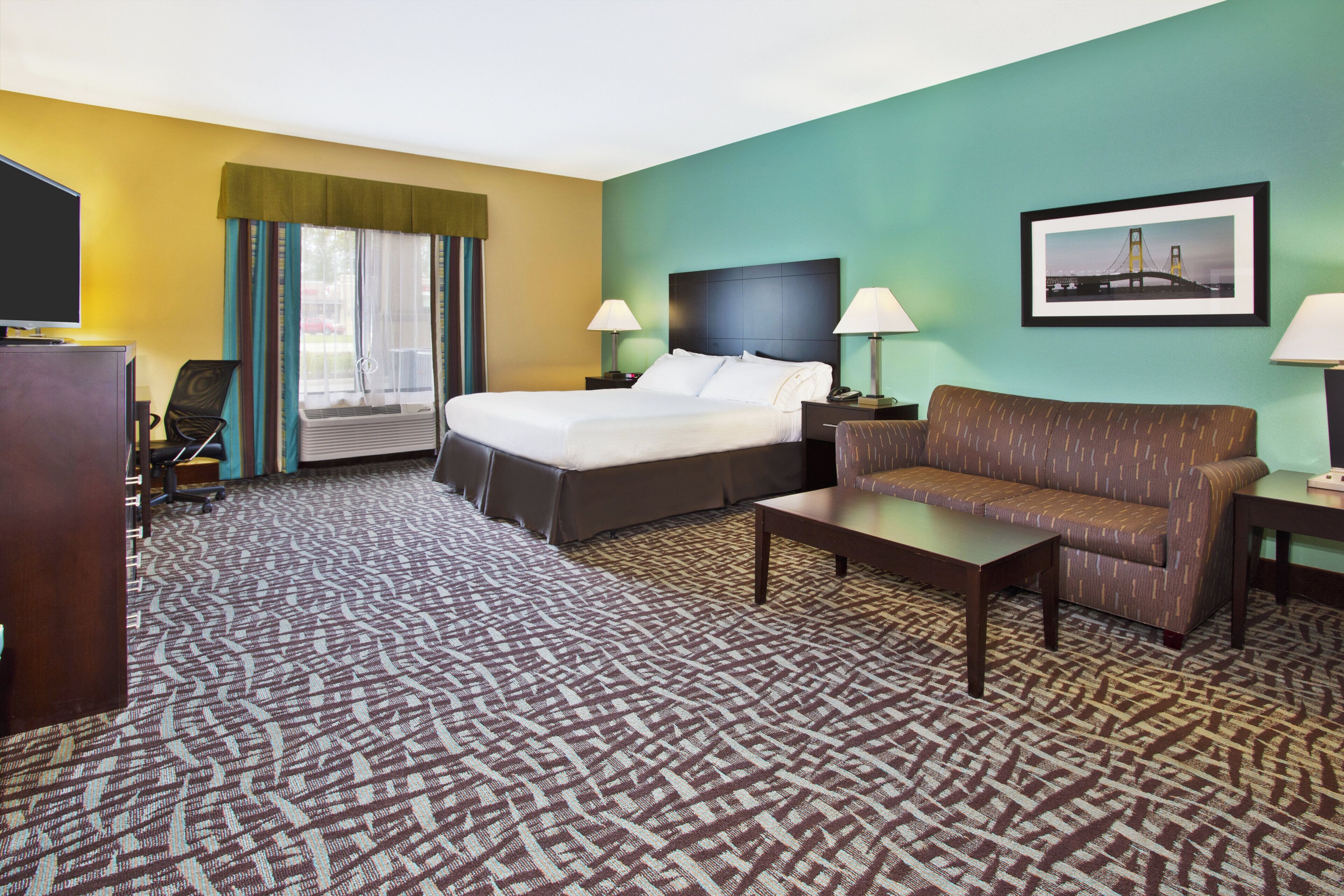 A hotel room with a bed, a sofa, a desk, a TV, and a framed picture on the wall.