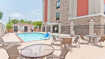 A pool area with lounge chairs and tables, a pool lift, and a building with a brick facade.