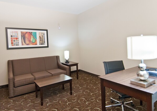 A hotel room with a brown patterned sofa, a small table with a lamp, a dining table with chairs, and a framed abstract artwork on the wall.