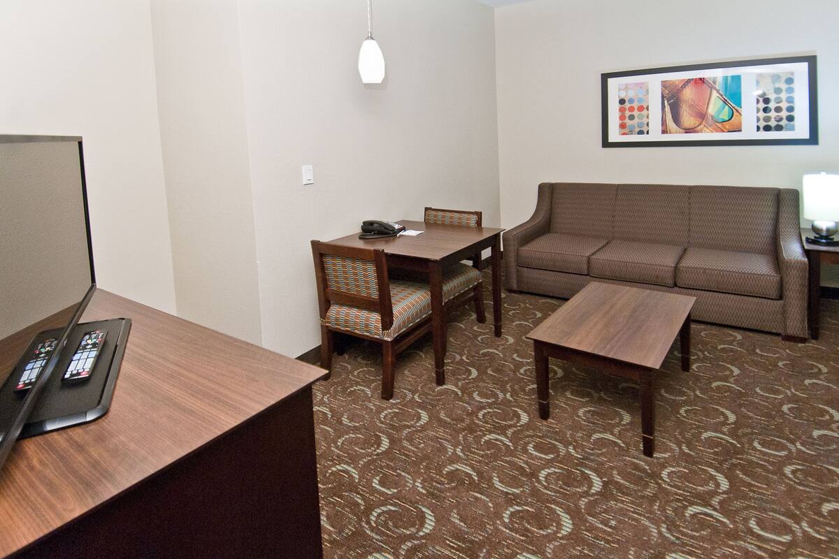 A hotel room with a wooden desk, a brown sofa, a dining table with chairs, a framed picture on the wall, and a patterned carpet.